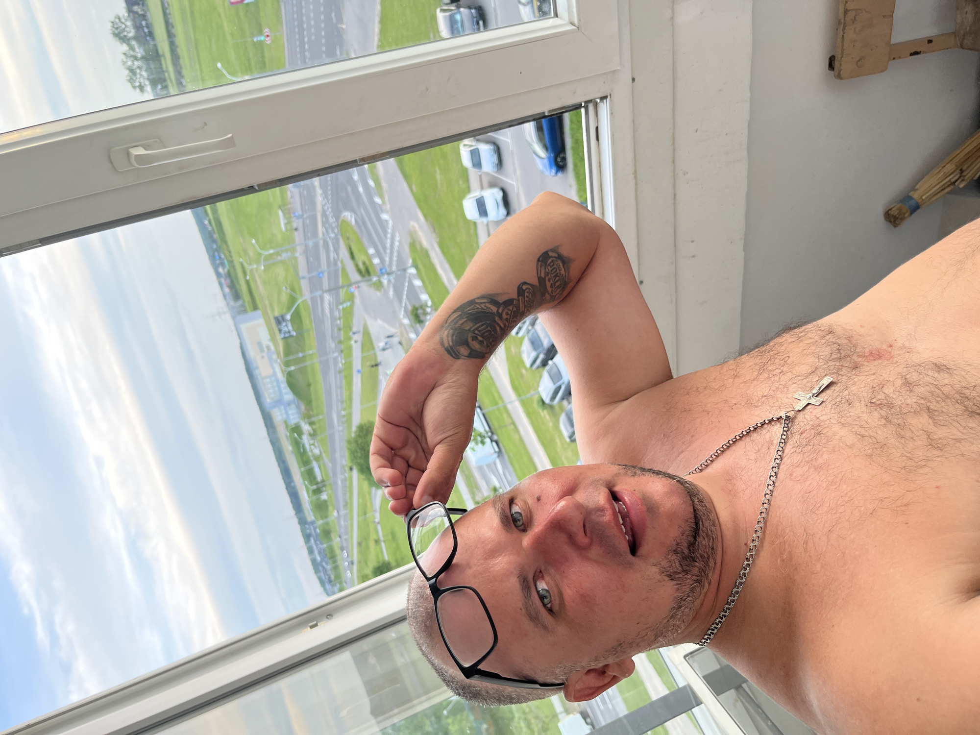 A shirtless man with a short beard and light-colored hair gazes directly into the camera, holding black-framed glasses near his forehead with his right hand. He wears a silver chain with a crucifix pendant around his neck, and a tattoo featuring poker chips and the word "POKER" is visible on his right arm. The man's chest is hairy, and his eyes are blue-green. He is positioned indoors, by a large window, which reveals a high-angle view of an urban landscape under a bright, partly cloudy daytime sky. The outdoor scene includes a wide multi-lane road with traffic circles, several cars, green verges, and distant buildings or land alongside a body of water. The image is rotated 90 degrees counter-clockwise. On the far right, a wooden structure and a broom are partially visible. This scene is set in Minsk, Belarus.