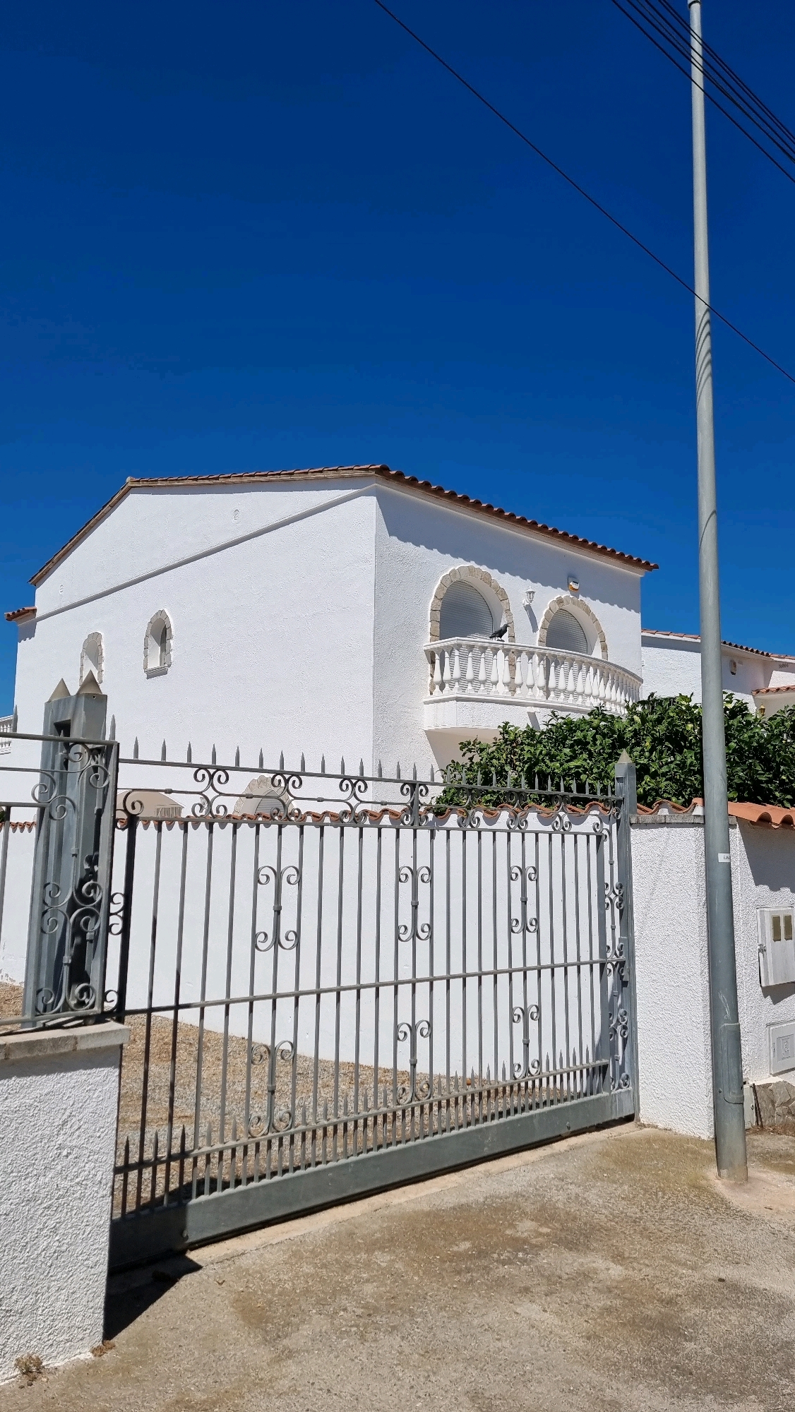 A white, stucco house with a tiled roof is seen from the exterior. The house features a balcony with decorative railing and two arched windows. A grey metal gate with ornate scrollwork is in the foreground, separating the viewer from the property. Behind the gate, a white wall with a tiled coping lines the front of the house. A grey metal utility pole stands to the right of the frame, with wires extending upwards and to the left. The sky is a clear, vibrant blue, and the ground in front of the gate appears to be a concrete or asphalt surface. The overall scene suggests a residential property located in Castelló d'Empúries, Spain, given the architectural style and the clear blue sky typical of the region.