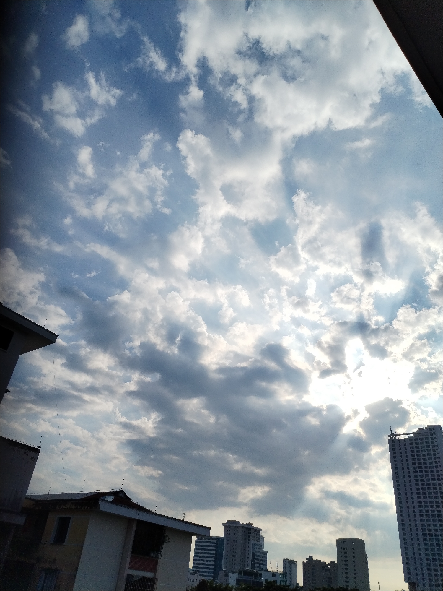 The image shows a view of the sky over Nha Trang Ward, Vietnam, with buildings visible in the foreground. The sky is predominantly blue with scattered white and gray clouds. Sunlight streams through the clouds, creating a bright, diffused light. Several buildings of varying heights are visible, including a tall, modern skyscraper on the right side of the frame. Other buildings appear to be older and of a lower profile. Overall, the image captures a dynamic sky and a partial urban landscape.