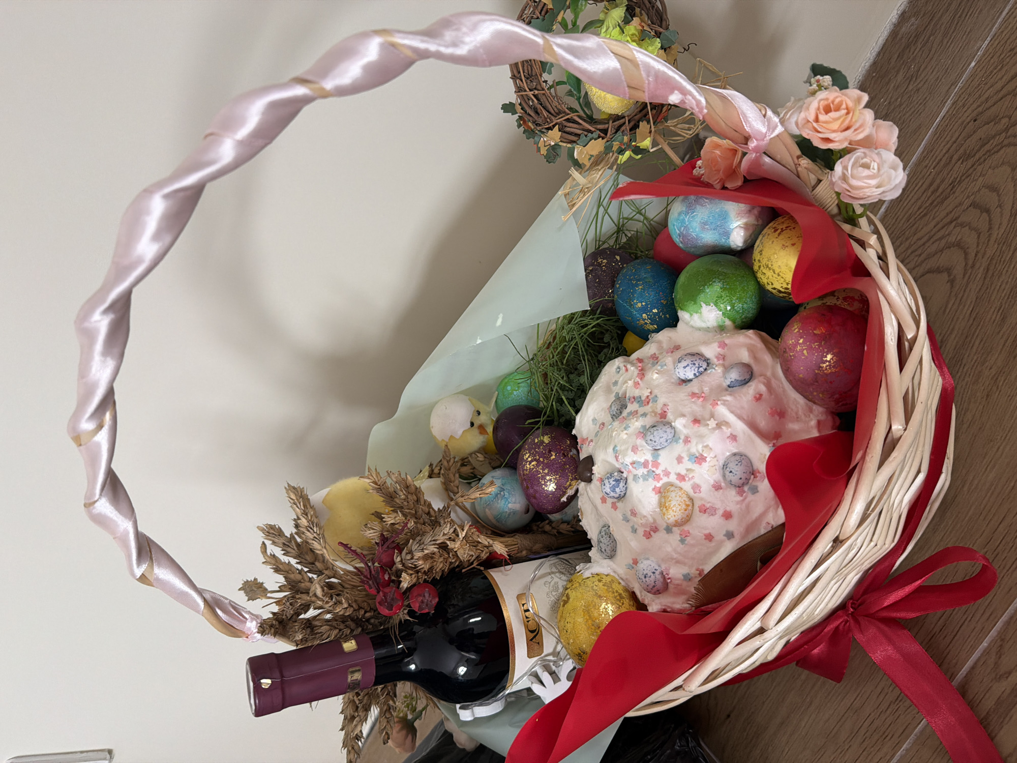 The image shows an Easter basket filled with various items. The basket is made of wicker and has a light pink ribbon woven around the handle. Inside the basket, there are decorated Easter eggs in various colors, a traditional Easter cake topped with frosting and decorative eggs, a bottle of wine, flowers, and dried wheat stalks. A red ribbon is tied to the side of the basket. The background includes a neutral-colored wall on one side and a wooden surface on the other. There is no evidence to suggest that the image was taken in the city of Krasnoyarsk, Russia.