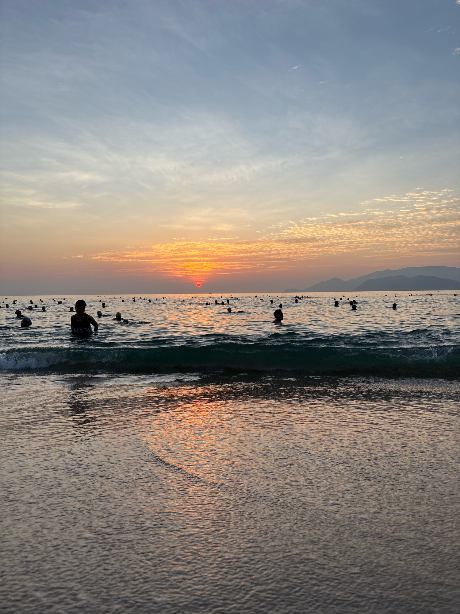 A group of people are swimming in the ocean near the coast of Nha Trang, Vietnam during sunset. The sky is filled with orange and pink clouds, and the sun is setting over the water. Mountains can be seen in the distance. The water is calm, with gentle waves lapping at the shore. Some people are in the shallow water near the beach, while others are further out in the ocean. A few individuals are visible in the foreground, wading in the water.