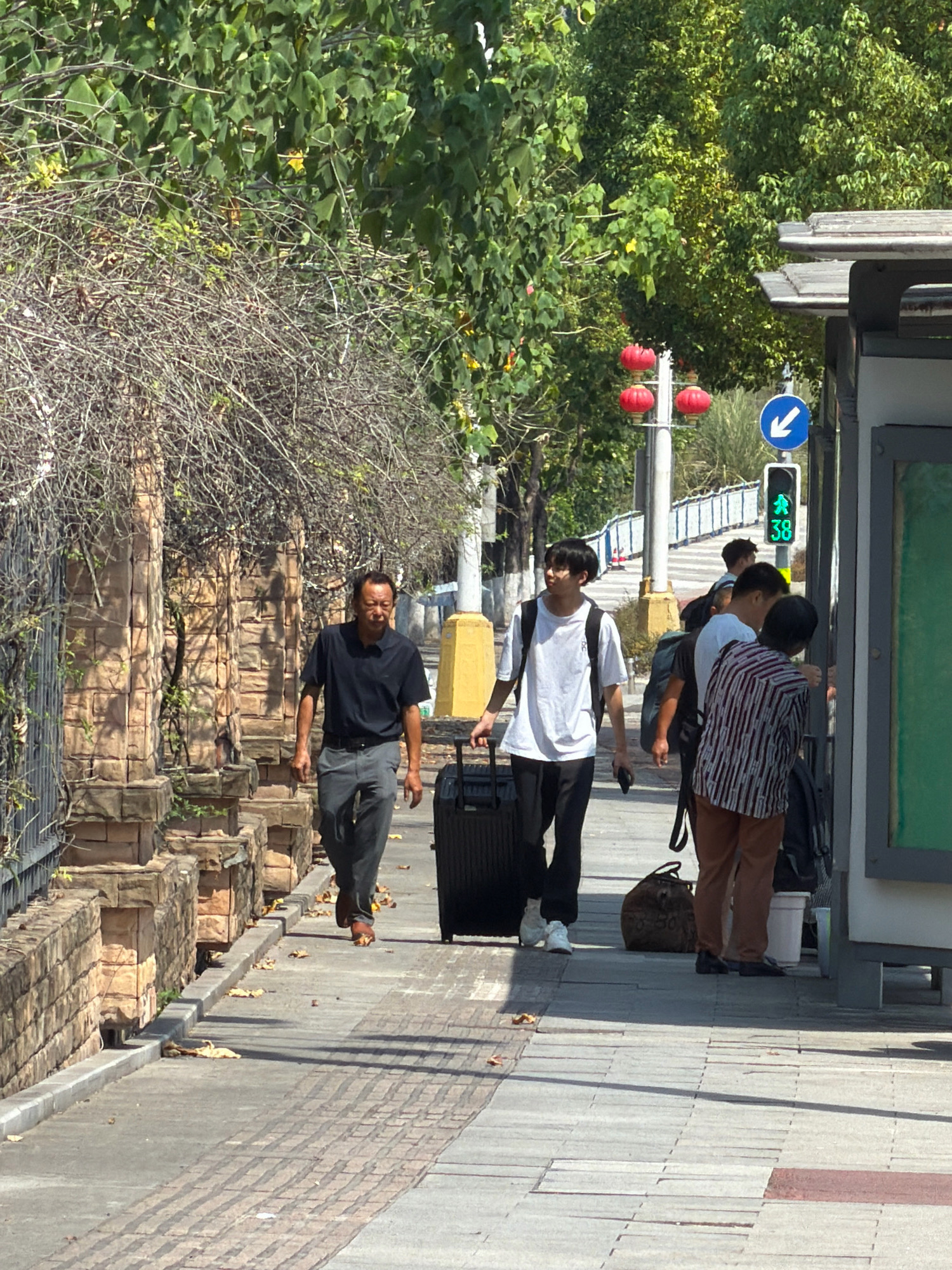 In Jiangjin, China, on a sunny day, two men and a younger person walk along a brick-patterned sidewalk next to a stone-embellished wall covered in dry branches. The man on the left, wearing a black polo shirt and grey pants, walks towards the camera, with a black rolling suitcase visible to his right. To his right, a younger person in a white t-shirt and black pants pulls a black rolling suitcase and looks to their left. Further to the right, at a bus stop shelter with a green display board, a third person in a striped shirt and brown pants is standing, facing away from the camera and reaching towards the shelter. A traffic light above them displays a green walking figure and the number "38". Red lanterns hang from a pole in the background, and lush green trees line the street.