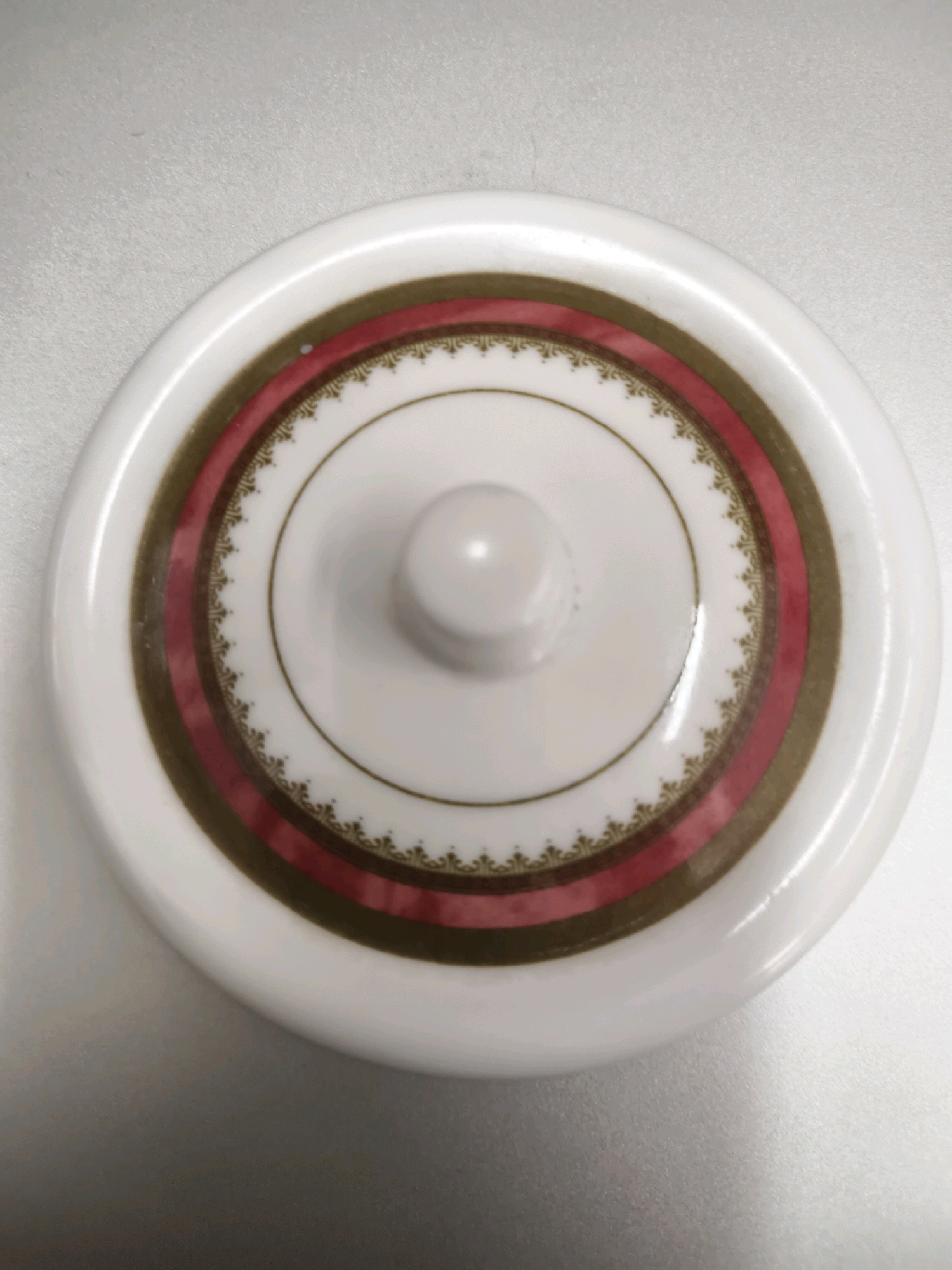 The image shows a close-up, overhead view of a white ceramic lid with decorative bands. The lid has a central, rounded knob. The decorative bands consist of an inner gold circle, followed by a wider reddish-pink band, then a thin gold band with a repeating intricate pattern, and finally another gold circle. The lid is placed on a textured, light-colored surface, possibly a tabletop or countertop. The image is well-lit, with a slight shadow cast by the lid on the surface. There is no indication of the location within the city of Salira, Indonesia.