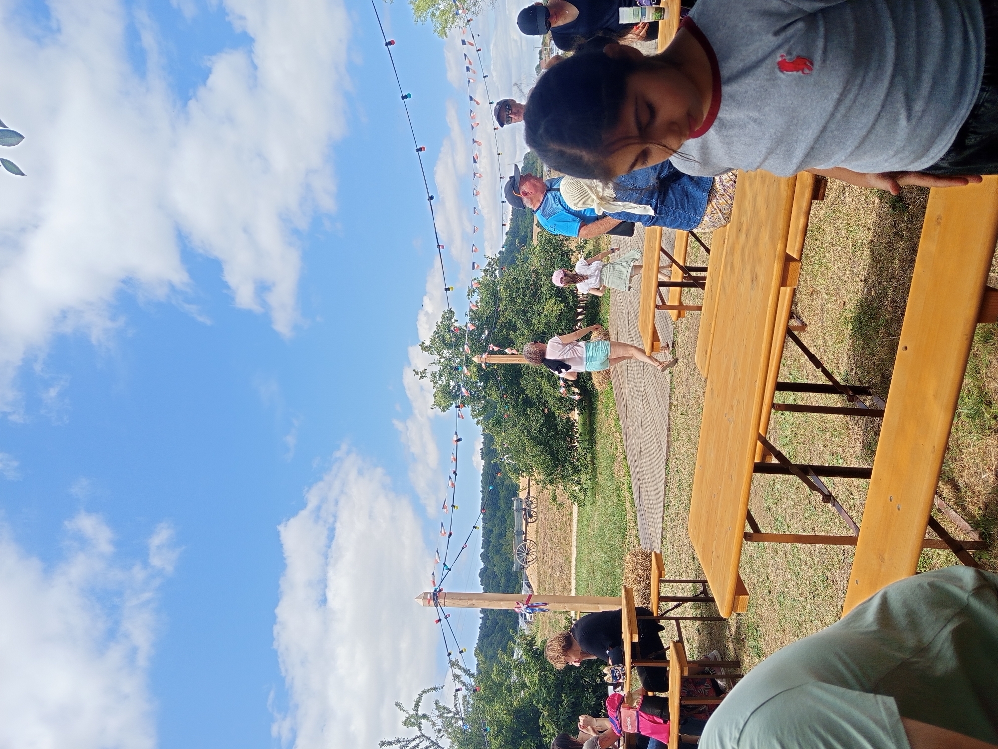 The image depicts an outdoor gathering in Azannes-et-Soumazannes, France, likely a local fair or festival, on a sunny day with scattered clouds. In the foreground, a young girl in a grey t-shirt with a red logo is looking down. To her left, a wooden picnic table and bench are visible, partially shaded. Further back, other people are seated at picnic tables and walking around. The scene includes trees, a wooden walkway, and a decorative string of colorful lights and small flags stretched between poles. In the background, a vintage wagon is parked on a dry, grassy area, with rolling hills covered in trees in the distance. The overall atmosphere is casual and festive.