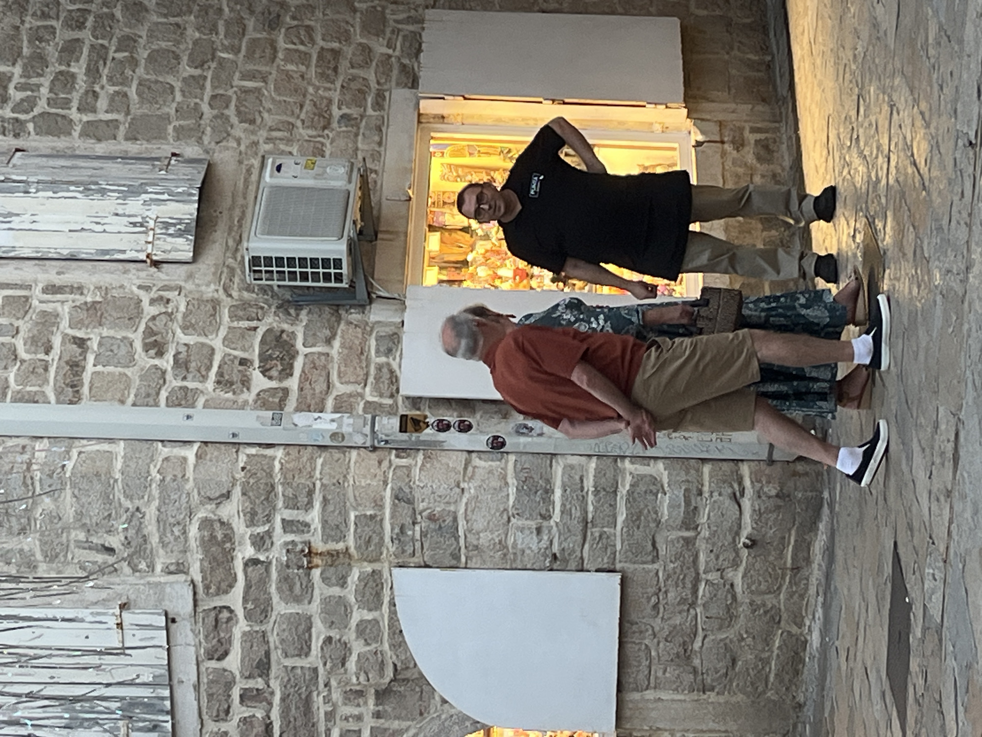 In Budva, Montenegro, at street level, three individuals are interacting outside a storefront. The building is constructed of rough-hewn stone blocks, with a weathered wooden shutter on the left and a modern air conditioning unit mounted on the wall above the shop. To the right, a storefront window is brightly lit, displaying various items inside, with white shutters on either side.
In the foreground, a man wearing an orange t-shirt and tan shorts stands with his back to the viewer, his hands clasped behind him. Beside him, a woman in a patterned dress, partially obscured, faces the storefront. To the right, a man wearing a black polo shirt with a logo and light-colored pants stands in the doorway of the shop, looking towards the other two individuals. He has glasses and his right hand rests on his hip. The ground is paved with irregular stone slabs, with reflections of the warm light from the storefront visible on the surface. The scene appears to be illuminated by the store's interior lighting and possibly ambient twilight.