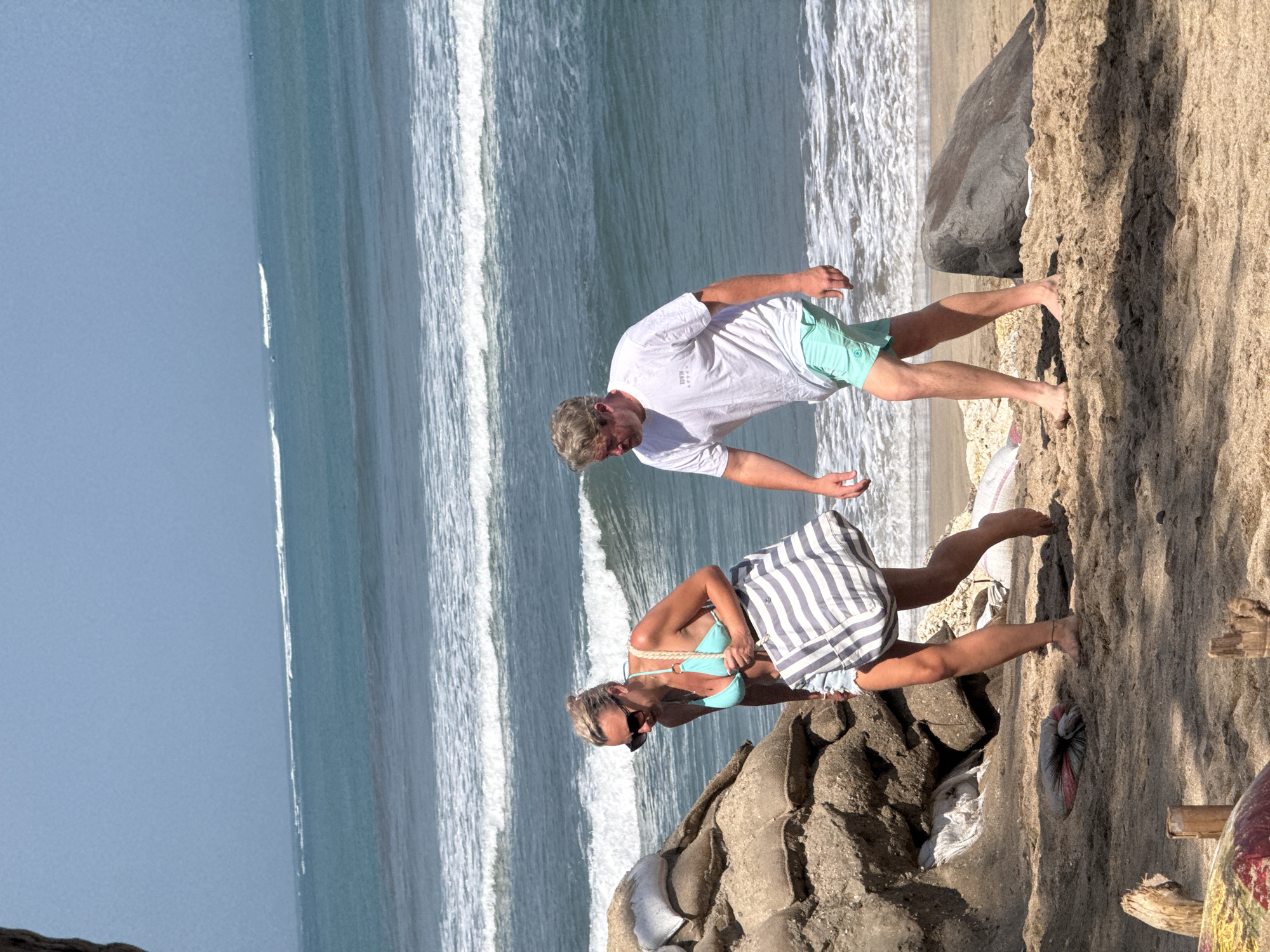 A man and a woman are walking on a sandy beach in Kuta, Indonesia. The woman is wearing a turquoise bikini and carrying a striped beach bag. The man is wearing a white t-shirt and teal swim trunks. In the background, the ocean has several lines of waves breaking. To the left of the woman is a large pile of sandbags. The sand on the beach is light brown, and there are some scattered rocks and driftwood. The sky is a clear blue.