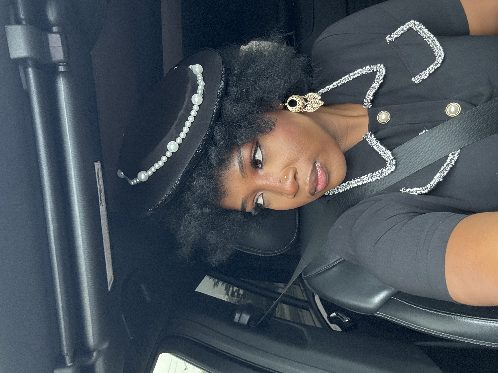 The image shows a woman sitting in the driver's seat of a car. She is wearing a black outfit with pearl accents and a black hat with pearls. She has gold earrings and a nose piercing. A seatbelt is buckled across her chest. The car interior is black leather. The photo appears to be taken indoors, within a car. There is a blurry window visible outside, suggesting an urban or suburban setting. The specific location in Lekki, Nigeria, cannot be determined from the image alone.