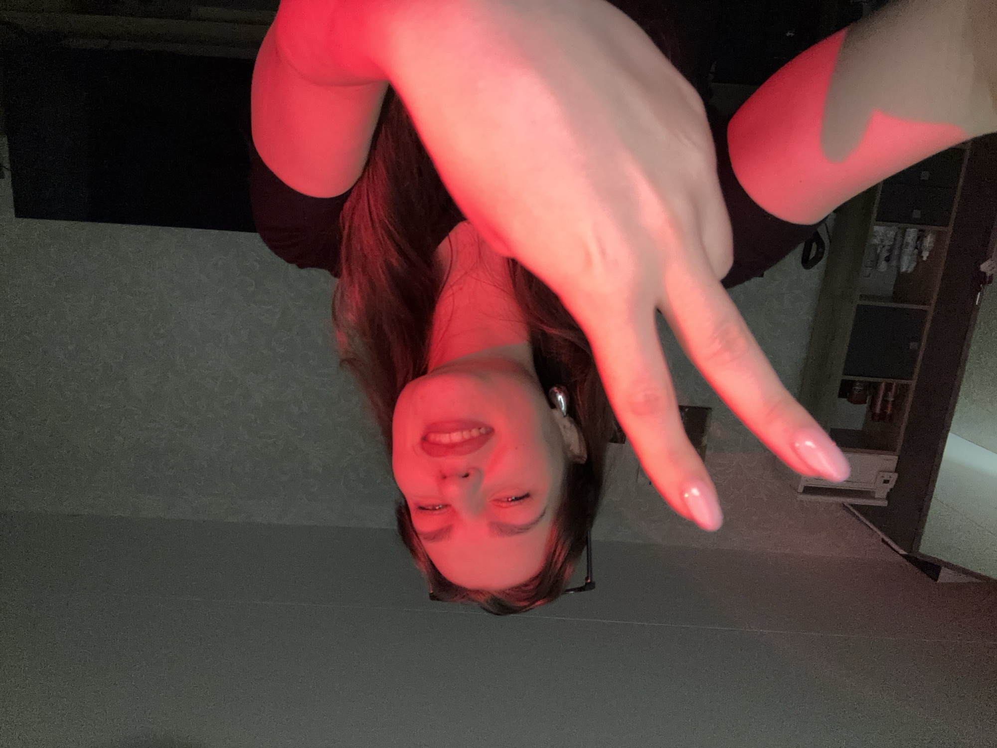 A young woman is pictured indoors, possibly in a residential setting, with red lighting casting a strong glow over her face and arm. She is looking towards the camera and smiling, with her right hand raised in a peace sign. Her fingernails are painted a light pink color. She is wearing a black t-shirt. In the background, to the left, a shelf unit with various items is visible. To the right, a dark television screen is present. The overall environment appears to be dimly lit, with the red light creating a dramatic effect. The image does not provide enough information to determine the specific location within the city of Chaikovsky, Russia.