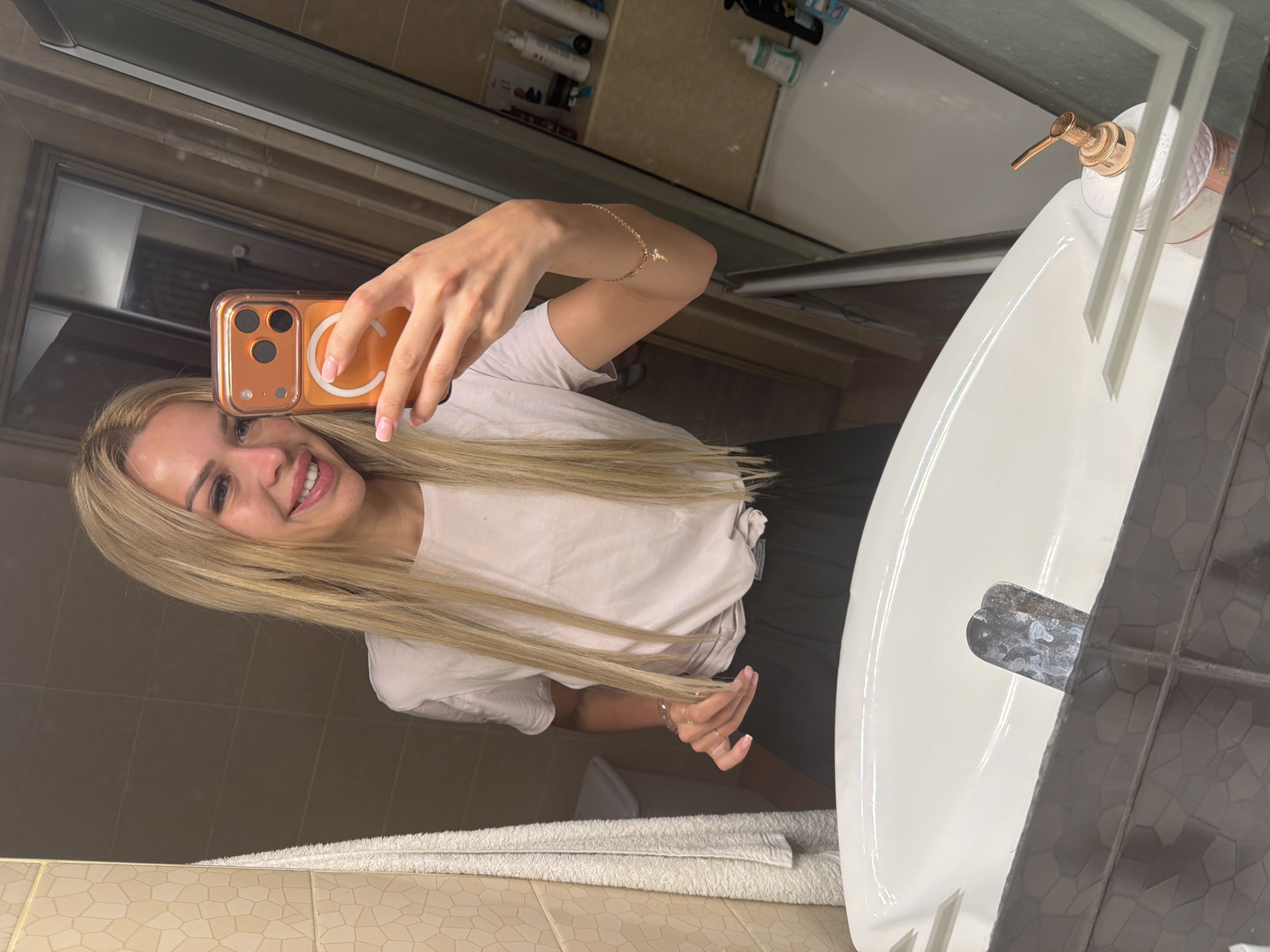 A young woman with long blonde hair and fair skin smiles at the camera while taking a mirror selfie. She is wearing a light-colored t-shirt and black pants, and she has on minimal makeup. She is holding her phone in her right hand, and a gold bracelet with a charm is visible on her left wrist. The background appears to be a bathroom, with a tiled wall, a sink, and a mirror reflecting more of the room. There are various toiletries visible on a shelf in the reflection. The image seems to have been taken indoors, likely in a residential setting.