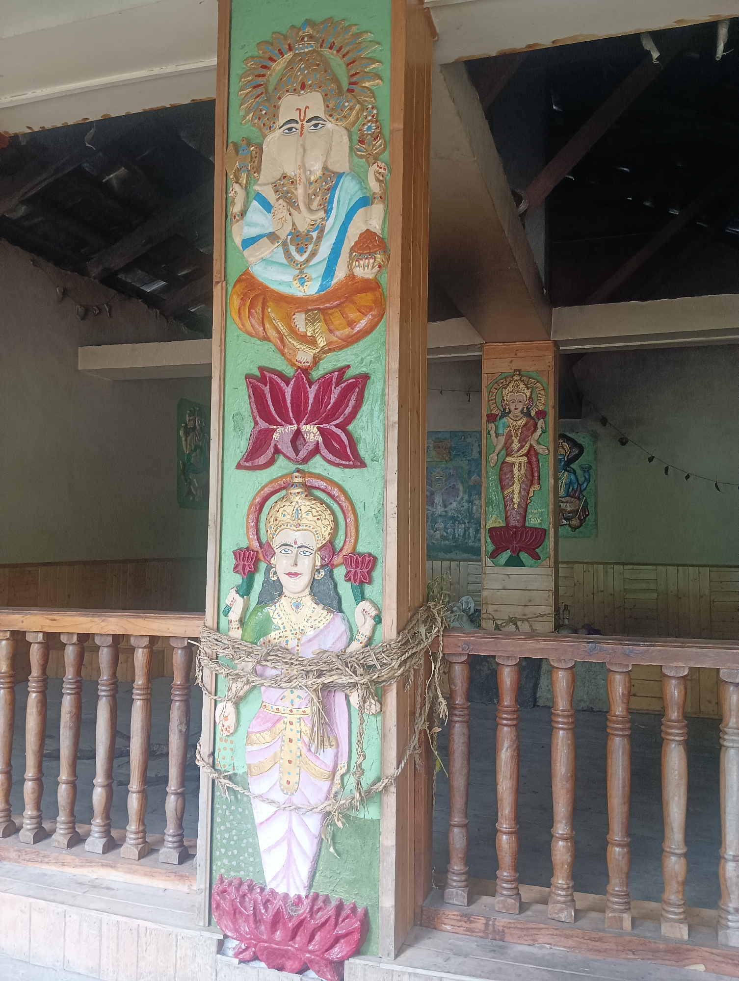 The image depicts an outdoor scene with intricate carvings of Hindu deities on a wooden pillar. The pillar is painted green and features a carving of Ganesha at the top, followed by a lotus flower, and then Lakshmi. The figures are brightly colored with gold, blue, pink, and orange accents. Ropes made of natural fibers are wrapped around the pillar, securing a bundle of dried grass.
In the background, another wooden pillar displays a carving of a standing female deity, possibly also Lakshmi, in a red sari. To the right of this pillar, a carving of a blue-skinned deity is visible. The background shows a rustic structure with a wooden railing in the foreground, suggesting an open-air pavilion or porch. Wooden beams and a corrugated roof are visible overhead. The environment appears to be in a state of some disrepair, with exposed beams and a somewhat unfinished appearance. The location is identified as Majhach, India, suggesting a cultural or religious site.