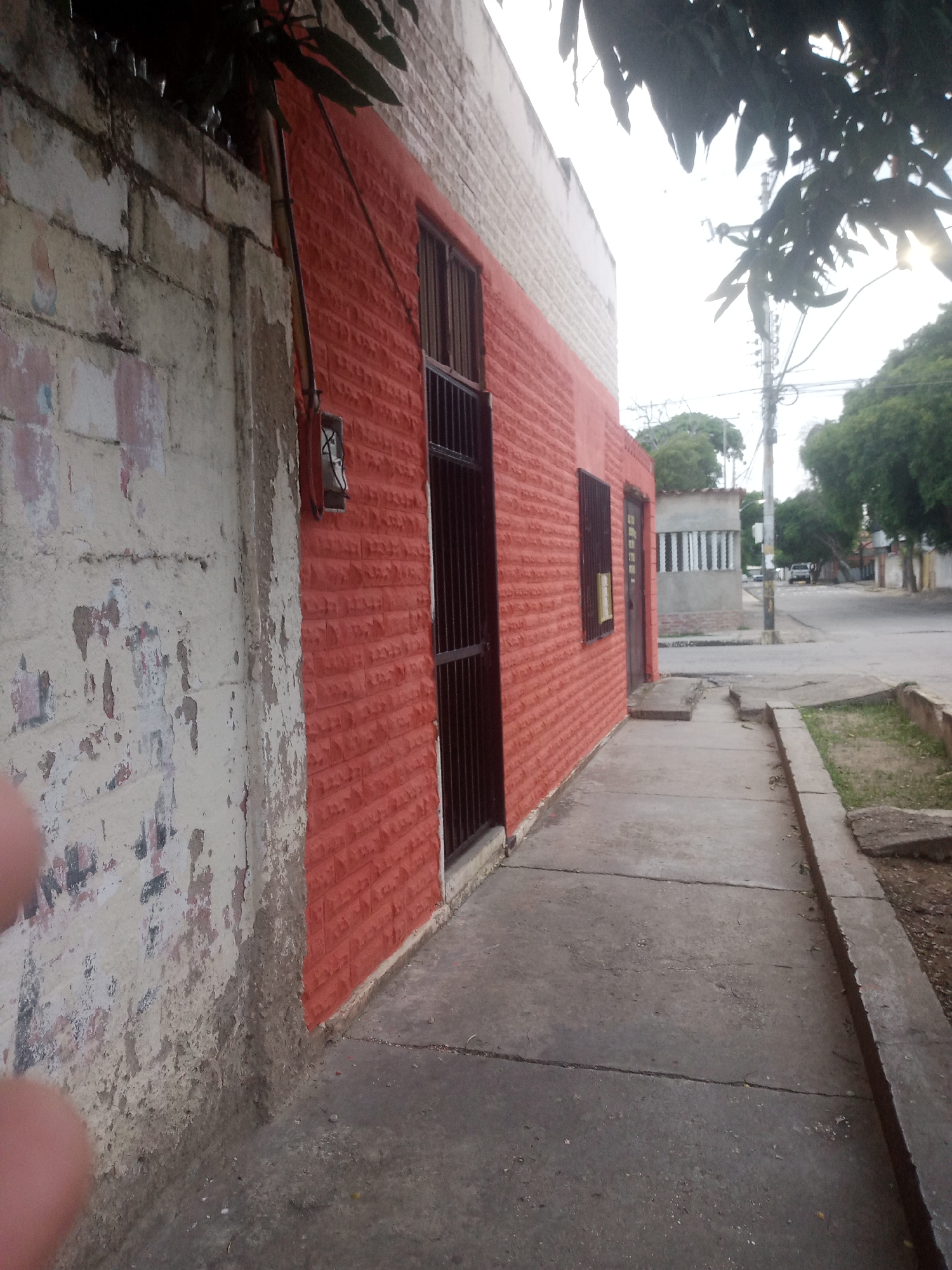 The image depicts a side view of a building with a red brick facade and a white upper section. A sidewalk runs along the side of the building, which appears to be in a street setting in the city of Santa Ana, Venezuela. To the left of the red building, there is a weathered, light-colored concrete wall with peeling paint and some graffiti. The red building has barred windows and a dark brown door. Further down the street, other buildings and trees are visible, as well as a utility pole. The sky is overcast.