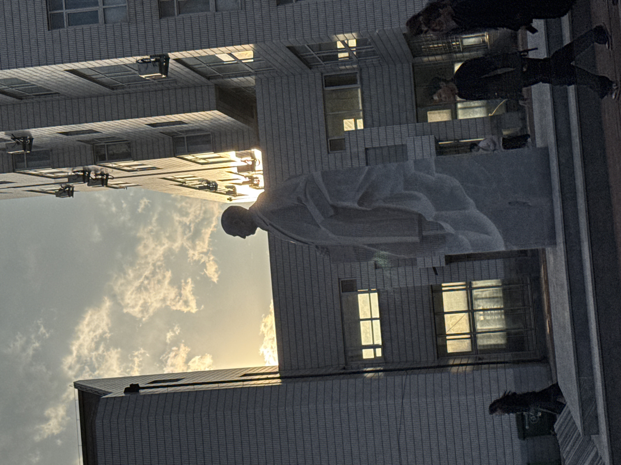 A low-angle shot depicts a statue of a bald-headed man in flowing robes, positioned in front of a multi-story building in Zhongtun, China. The building's architecture features numerous windows. The sky is visible in the background. Several people are walking on the sidewalk.
