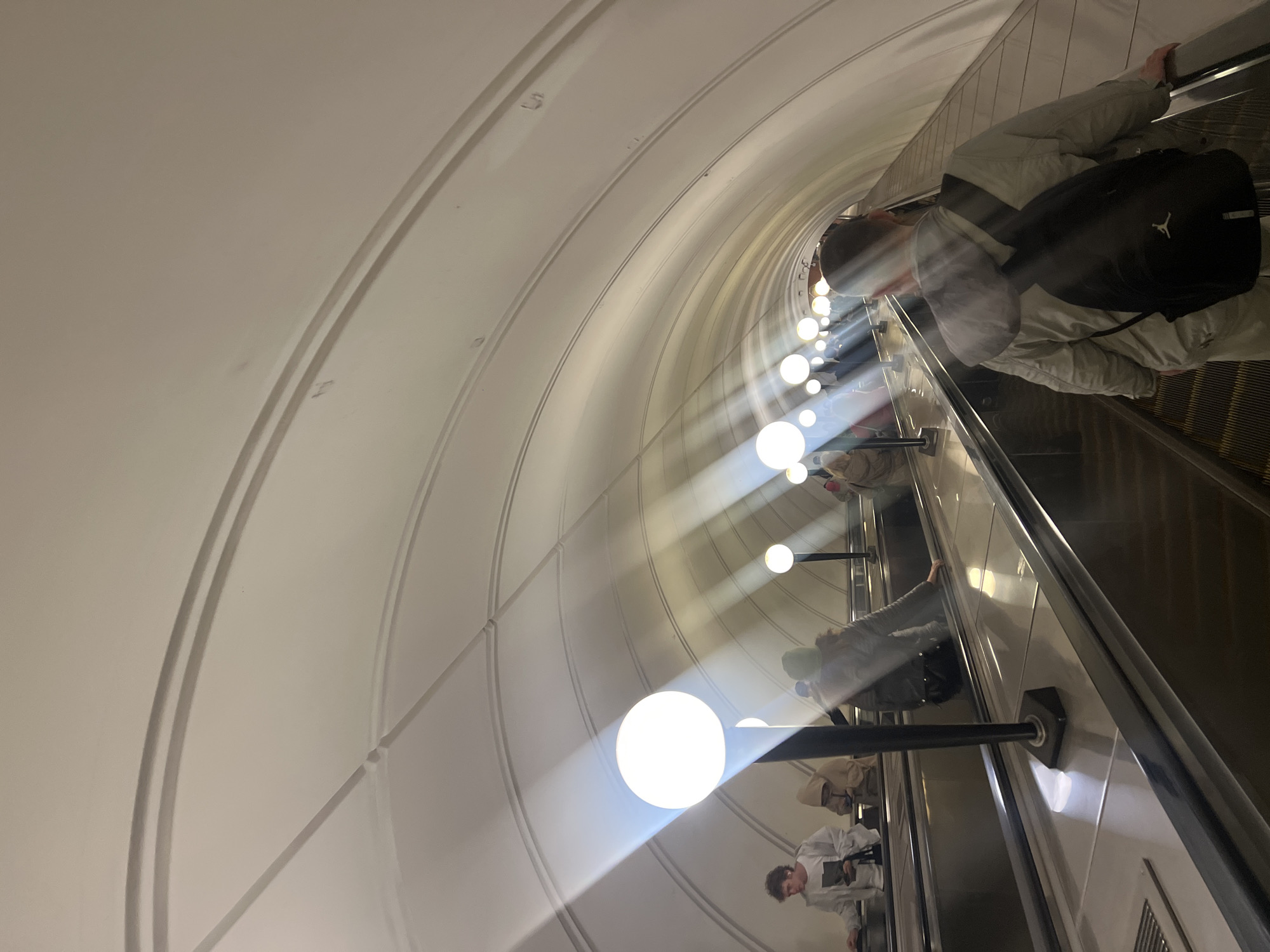The image shows an escalator ascending in the Moscow Metro. The tunnel is arched and illuminated by a series of spherical lights. People ride the escalator. One person wears a backpack and jacket. The environment is clean and modern.