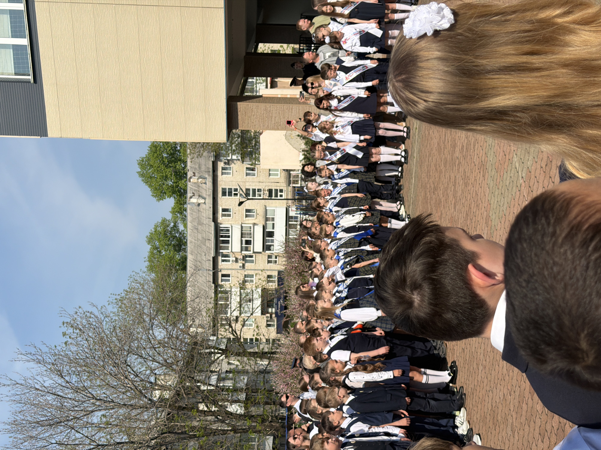 A large group of students in school uniforms is standing in rows outdoors. The students are wearing white blouses, dark skirts or pants, and many have white ribbons or bows in their hair. They appear to be participating in a school event or ceremony. The location seems to be Birobidzhan, Russia.
Behind the students are buildings, including a multi-story apartment building and part of a school building. A tree with bare branches is in the foreground.