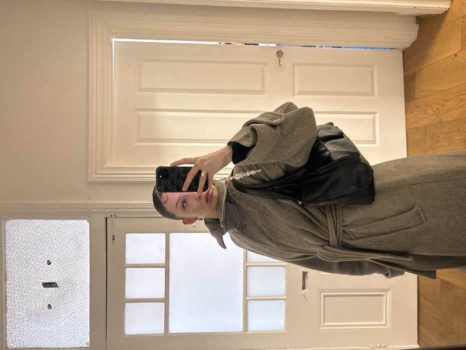 A mirror selfie depicts an individual indoors, likely within a residence in London, United Kingdom, evidenced by the architectural style of the interior doors. The subject, visible from mid-thigh upwards, is wearing a long, light grey or beige wool wrap coat with a high standing collar, secured at the waist by a self-fabric belt. A black long-sleeved garment is visible beneath the coat sleeves. A black leather shoulder bag is carried on the subject's left shoulder. The subject's right hand holds a dark-cased smartphone with a geometric grid pattern, obscuring the majority of their face. A white and translucent beaded phone charm or strap extends from the phone. The left eye, nose with dual piercings (septum and nostril), and a silver-colored ring on the left hand's middle finger are discernible. The immediate environment consists of light-toned wooden floorboards and white painted walls. In the reflection, a white painted wooden door on the left features multiple small glass panes within its upper section, topped by a horizontal textured glass transom window. To the right, a second white painted wooden door stands ajar, revealing a narrow vertical glimpse of an adjacent room with a muted blue hue. The door and window styles are consistent with early 20th-century residential architecture prevalent in London.