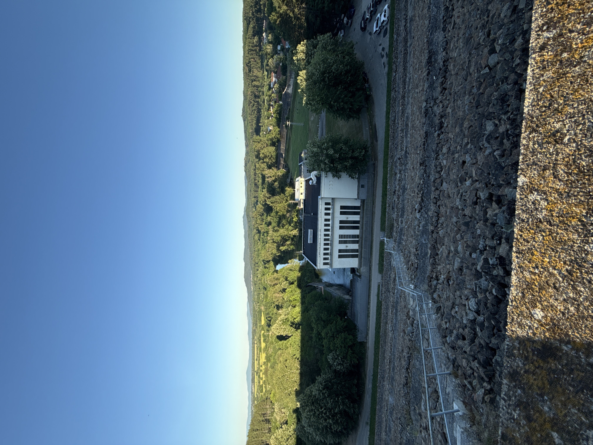 The image captures a wide shot of a dam and its surrounding landscape. In the foreground, a steep embankment made of large, dark rocks is visible, with a metal railing guiding a path down. The embankment transitions into a grassy area with a low fence, bordering a road where several cars are parked.
Dominating the mid-ground is a white, rectangular building with a dark roof, featuring rows of large windows. This structure appears to be part of the dam's operational infrastructure. Adjacent to it, a body of water flows, presumably released from the dam, and a concrete wall lines its bank.
The background reveals a lush green landscape with dense trees and rolling hills. A river or stream meanders through the trees, and a small settlement with houses and a church can be seen nestled amongst the foliage. The sky is clear and bright blue, indicating fair weather. The overall scene suggests a tranquil, natural setting with industrial elements integrated into the environment. The location is identified as the dam of Koprinka, Bulgaria.
