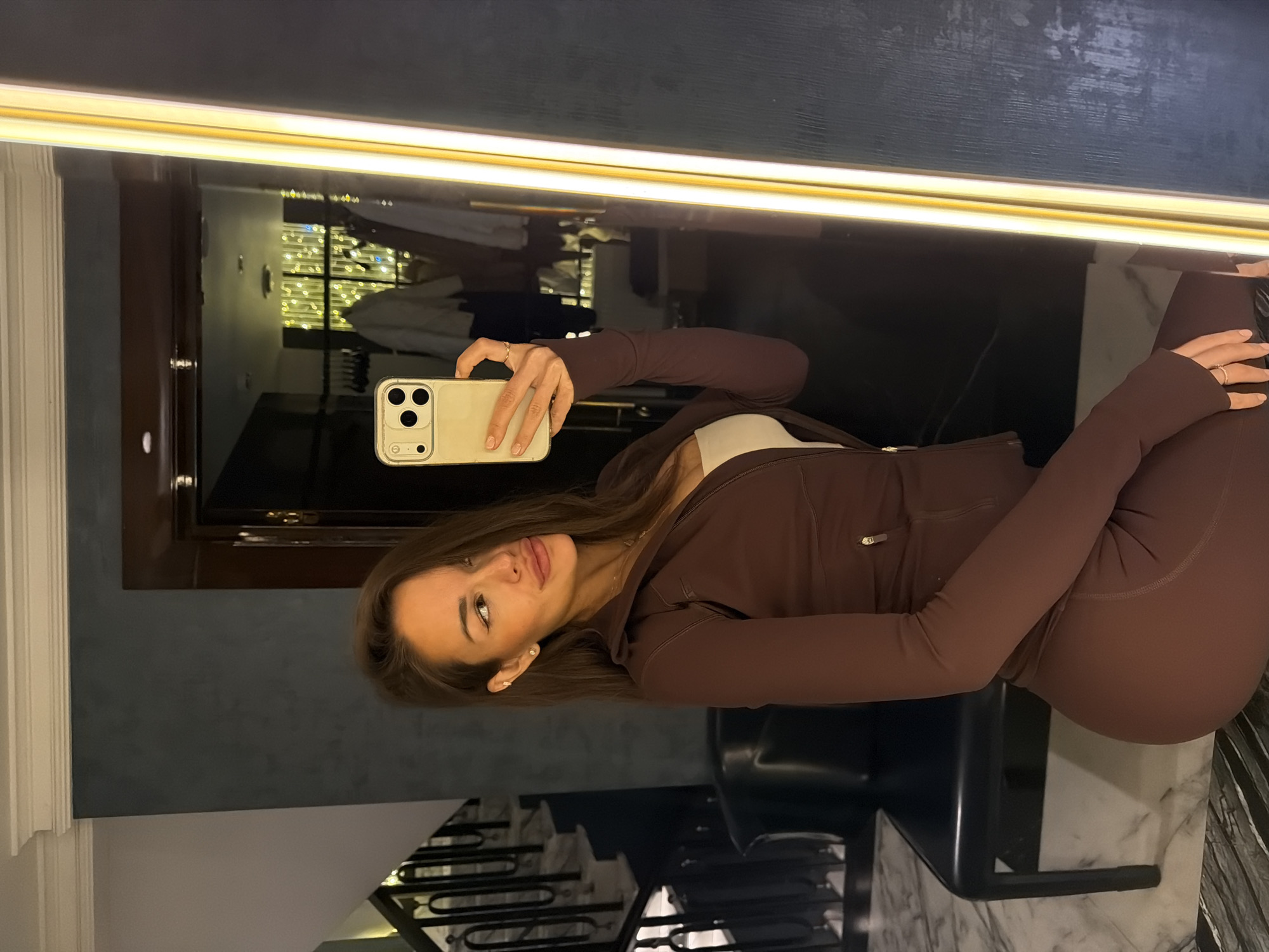 A young woman with long brown hair takes a mirror selfie in what appears to be a hotel room. She is wearing a brown athleisure set, consisting of a zip-up jacket and leggings, with a white crop top underneath. The woman is looking at her phone, which she holds in her right hand. Her left hand is resting on her knee. The background shows a staircase with a marble floor and dark railings, a dark leather bench, and a large mirror with a vertical LED light strip. In the reflection of the mirror, there are clothes hanging on a rack and decorative lights. The overall ambiance suggests a luxurious, possibly a hotel, setting in Moscow, Russia.