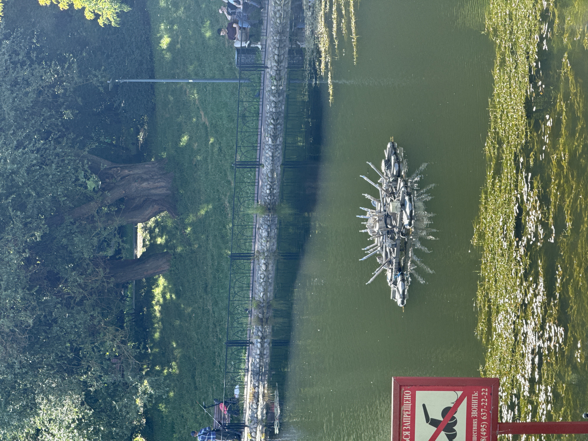 The image depicts a serene lake in a park, likely in Moscow, Russia, on a bright, sunny day. A man in a plaid shirt is fishing on the bank of the lake, with a red backpack and a fishing rod. Several people are sitting on a bench in the background, enjoying the view. In the center of the lake, a unique floating device with numerous metallic parts is visible. The foreground features a red sign with a graphic symbol of a person swimming, crossed out, indicating swimming is prohibited. Below this, text in Russian provides a phone number for emergencies. The lake is surrounded by lush green grass and mature trees, casting dappled shadows on the ground, creating a peaceful natural setting. The water is calm, reflecting the surrounding greenery and sky.