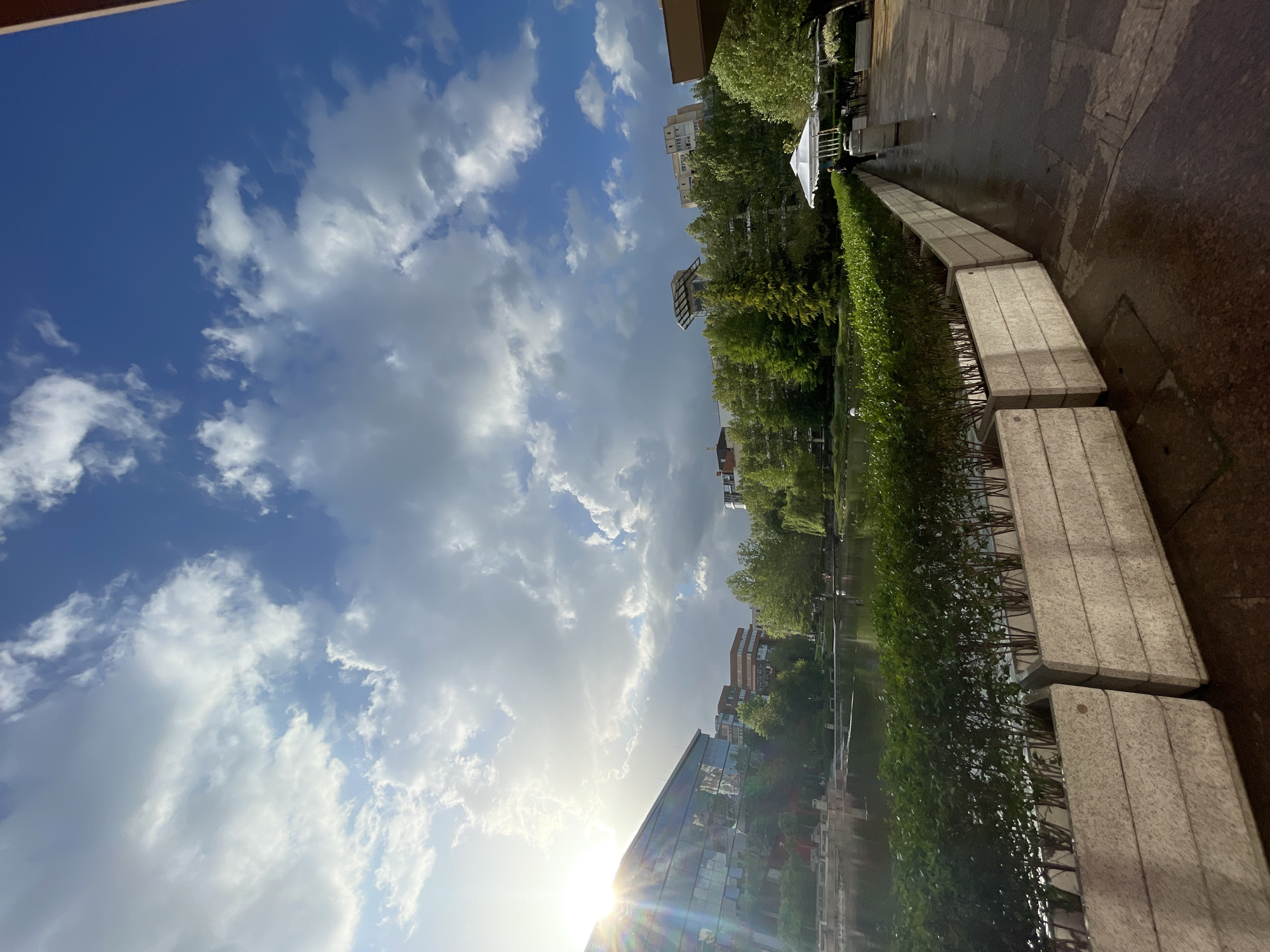 The image depicts a park scene in Sofia, Bulgaria. A canal runs through the park, bordered by a hedge and a series of concrete blocks. The backdrop includes buildings, mostly with leafy trees that line the parkway. The sky is mostly visible and contains a variety of cumulus clouds, with the sun visible over the horizon. The scene is bathed in sunlight, indicating the photo was taken near sunrise. A wet walkway is on the right side of the image. The building to the lower left looks like a modern glass structure.