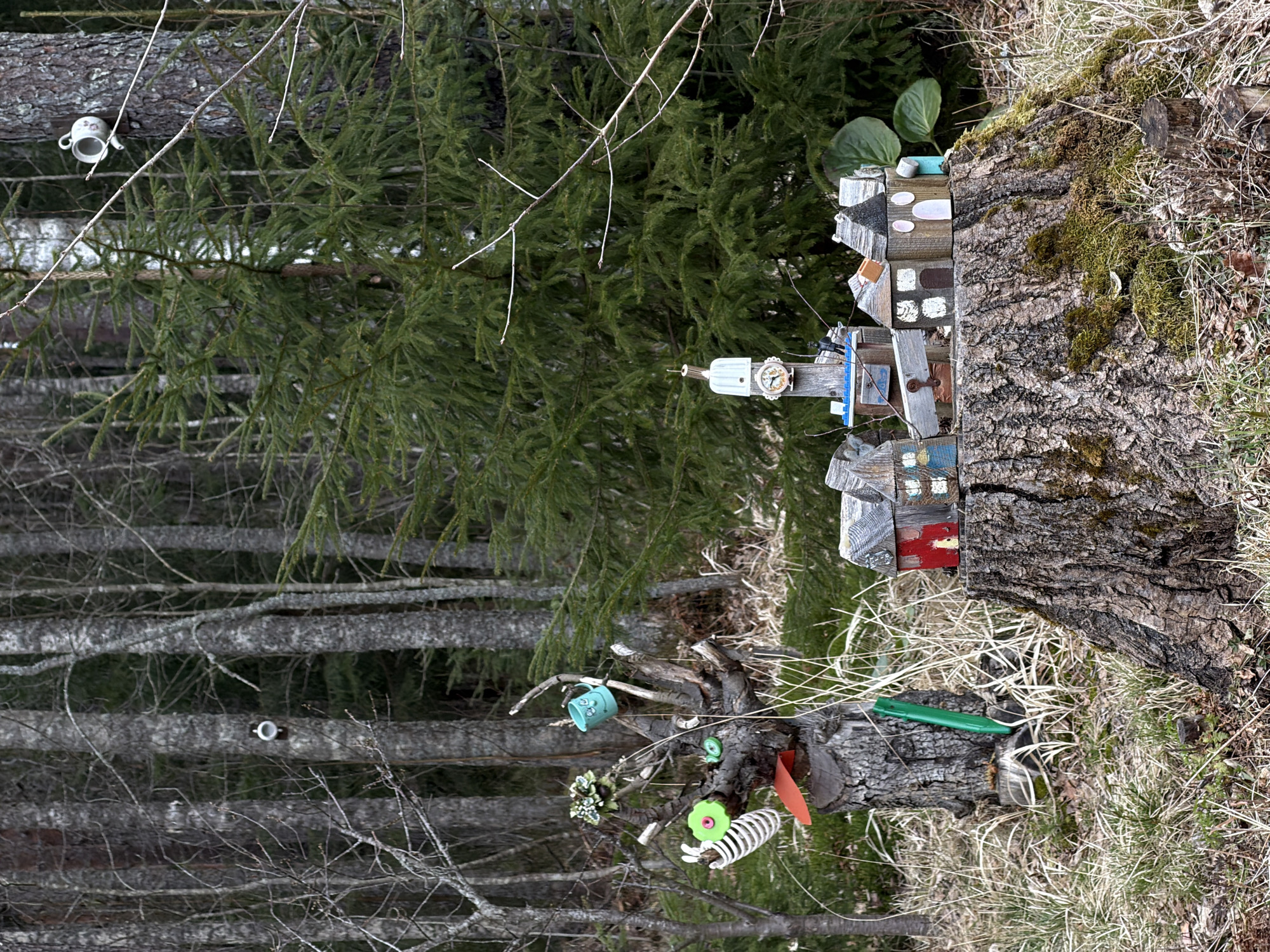 The image shows a forested area, possibly in Glubokoe, Russia, based on the prompt. A tree stump dominates the foreground, adorned with a miniature village of small, wooden buildings. The houses are varied in design and color, creating a whimsical effect. To the left of the stump and attached to the base, found objects like miniature teacups and plastic toys are affixed to the tree. Behind the stump is a dense forest of thin trees with bare branches, and another evergreen tree. The environment is natural with grass and debris around the tree stump.