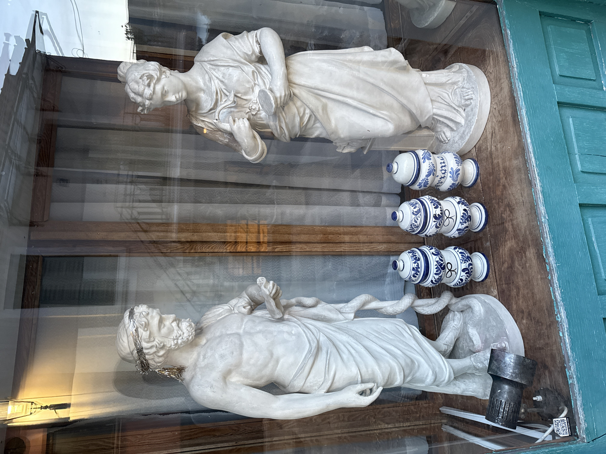 A shop window display in Herceg Novi, Montenegro, features two white marble statues, one male and one female, lying horizontally. Between the statues are three blue and white decorative pottery pieces, arranged vertically. The statues and pottery are resting on a wooden shelf behind a glass window. Reflections from the street are visible on the glass. A section of blue wooden shutters is visible along the right edge of the window frame. An older-style light fixture sits behind the male statue.