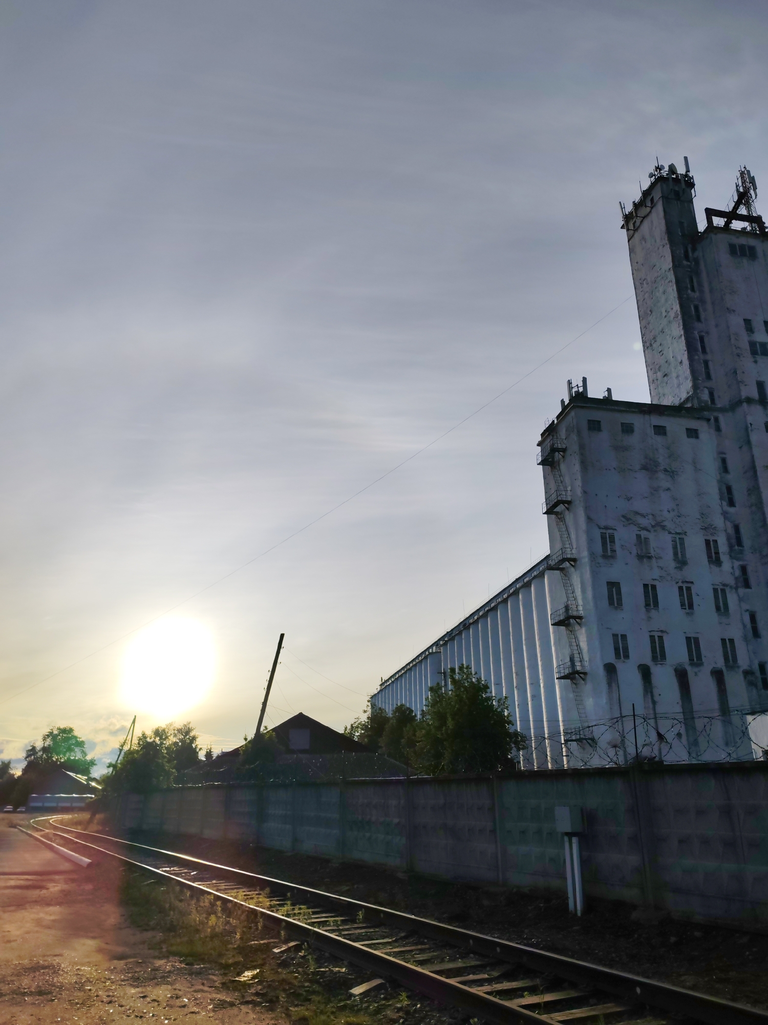 The photograph depicts a railway track curving toward the left foreground, adjacent to a concrete wall topped with barbed wire. Beyond the wall, trees obscure a single-story building with a gabled roof. To the right, a large, white, multi-story industrial building, possibly a grain elevator, dominates the scene, with ladders visible on its side. Antennas are present on the top of the building. The sky is bright, indicating either sunrise or sunset, with the sun positioned on the left side of the image. The photograph was taken in Bol'shie Mkhi, Russia.