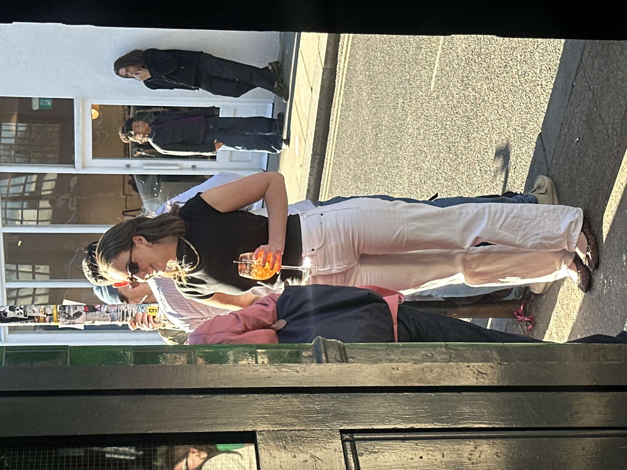 This image captures an outdoor urban scene, likely viewed from inside a building or through a window, as indicated by the dark wooden frame in the immediate foreground. Several people are depicted on a sunny day in what appears to be London.
In the foreground, a woman stands prominently, holding a glass of an orange-colored drink, possibly an Aperol Spritz, with ice and fruit garnishes. She wears sunglasses, a black top, a gold necklace, and distinctive white wide-leg trousers paired with leopard-print ballet flats. Behind her, partially obscured, is a man in a blue beanie, a striped shirt, and sunglasses. Another person in a pink top and dark trousers is visible further back in this group. To the left of this group, a pole or signpost is covered in various stickers, some featuring text fragments like "SMOKING", "FILM", and numbers such as "6" and "9".
In the mid-ground, two men lean against a white building with large windows reflecting parts of the street and sky. One man has dark hair and wears a dark jacket and trousers. The other, with curly hair, is dressed in a dark jacket and jeans. Visible through a window reflection in the background is a green sign with a white running man pictogram, typically indicating an exit.
The setting is an urban street or sidewalk, characterized by grey asphalt and paving stones. The bright sunlight casts long, distinct shadows on the ground, suggesting it is either late afternoon or early morning. The weather is clear and sunny. The overall activity appears to be casual socialising or waiting outdoors.