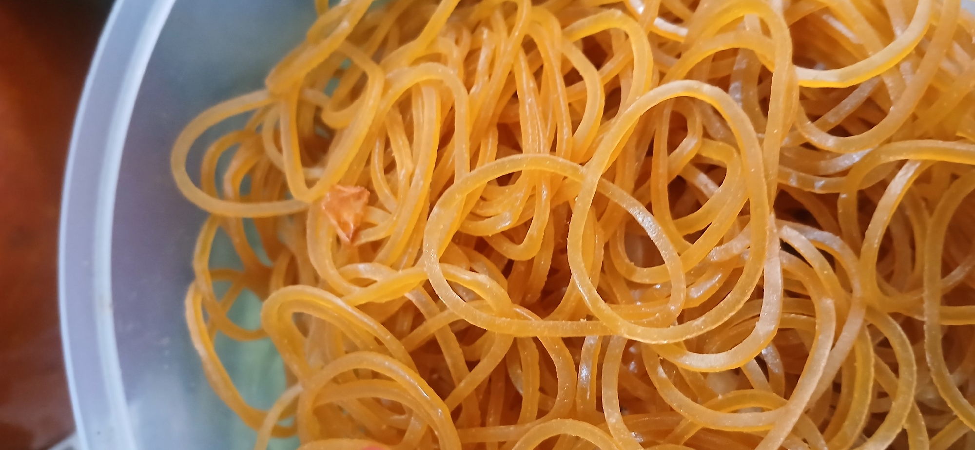 A close-up, overhead shot of a translucent plastic container filled with many yellow rubber bands. The container appears to be a light blue-grey color along the rim. The rubber bands are tightly packed, creating a textured, wavy pattern. There is a small, orange, triangular piece of what appears to be dried food or debris mixed in with the rubber bands. The background is blurred, with hints of brown and orange, suggesting other objects or surfaces. The scene is indoors and likely within a residential or storage area in West Jakarta, Indonesia.