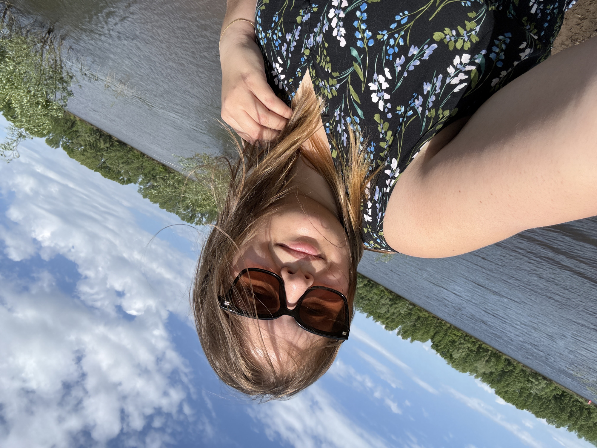A young, light-skinned woman with shoulder-length brown hair and sunglasses stands in front of a body of water. She wears a dark-colored floral dress and is holding her hair. The backdrop features a tree-lined shore and a partly cloudy sky. The image seems to be taken near water, possibly the Kama River. While precise determination of the location in Chaikovsky, Russia is difficult without additional context, the presence of the Kama River near the city suggests this could be a possible setting.