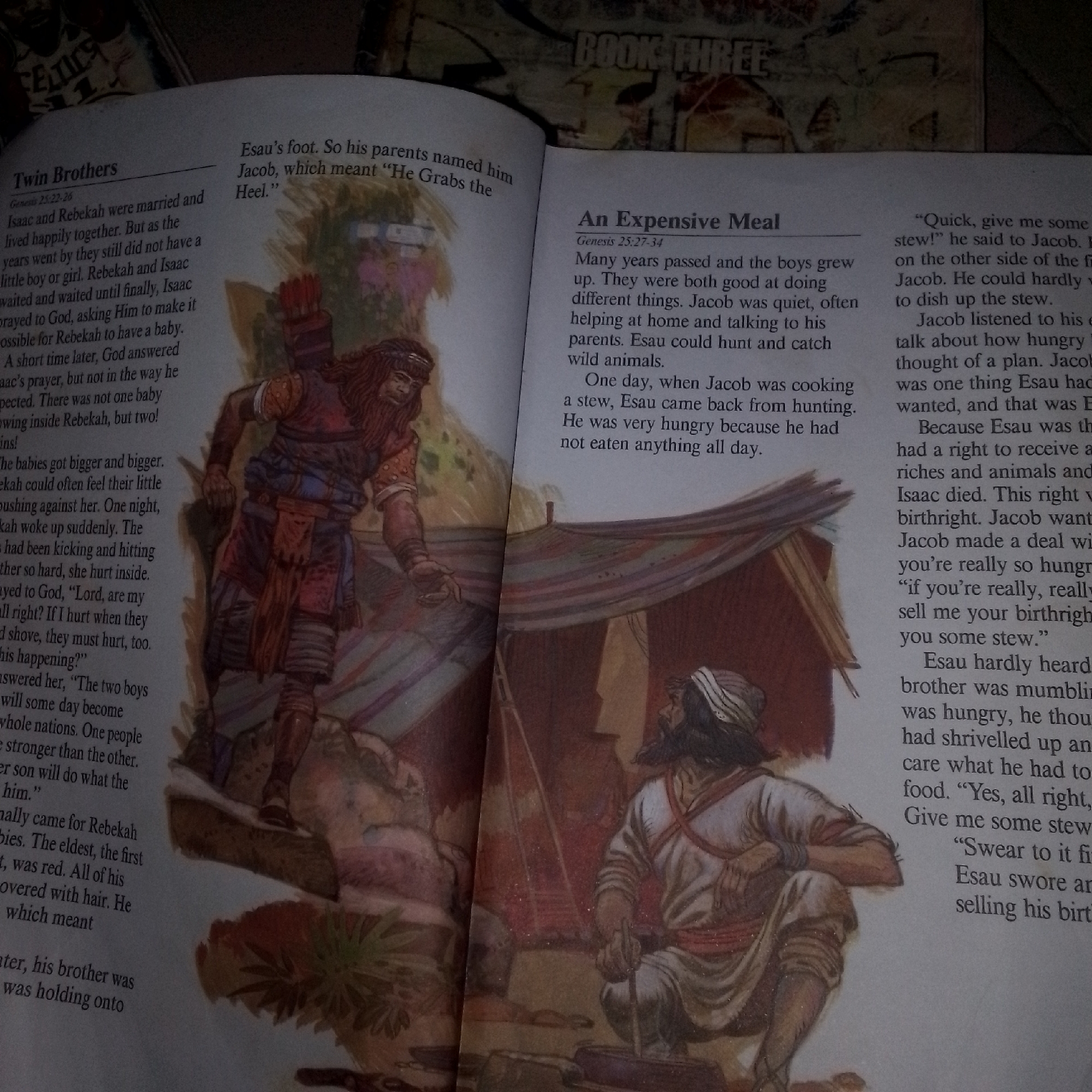 The image shows two open pages of a book. The left page has a title "Twin Brothers" and text below it, accompanied by an illustration of a man with a bow and arrow. The right page has a title "An Expensive Meal" and text, with an illustration of two men, one cooking over a fire outside a tent, and another man observing. The book appears to be a religious text, possibly a children's Bible, given the illustrations. The lighting in the image is dim, suggesting it might have been taken in low light conditions. There is no visible information indicating the location within Jalingo, Nigeria.