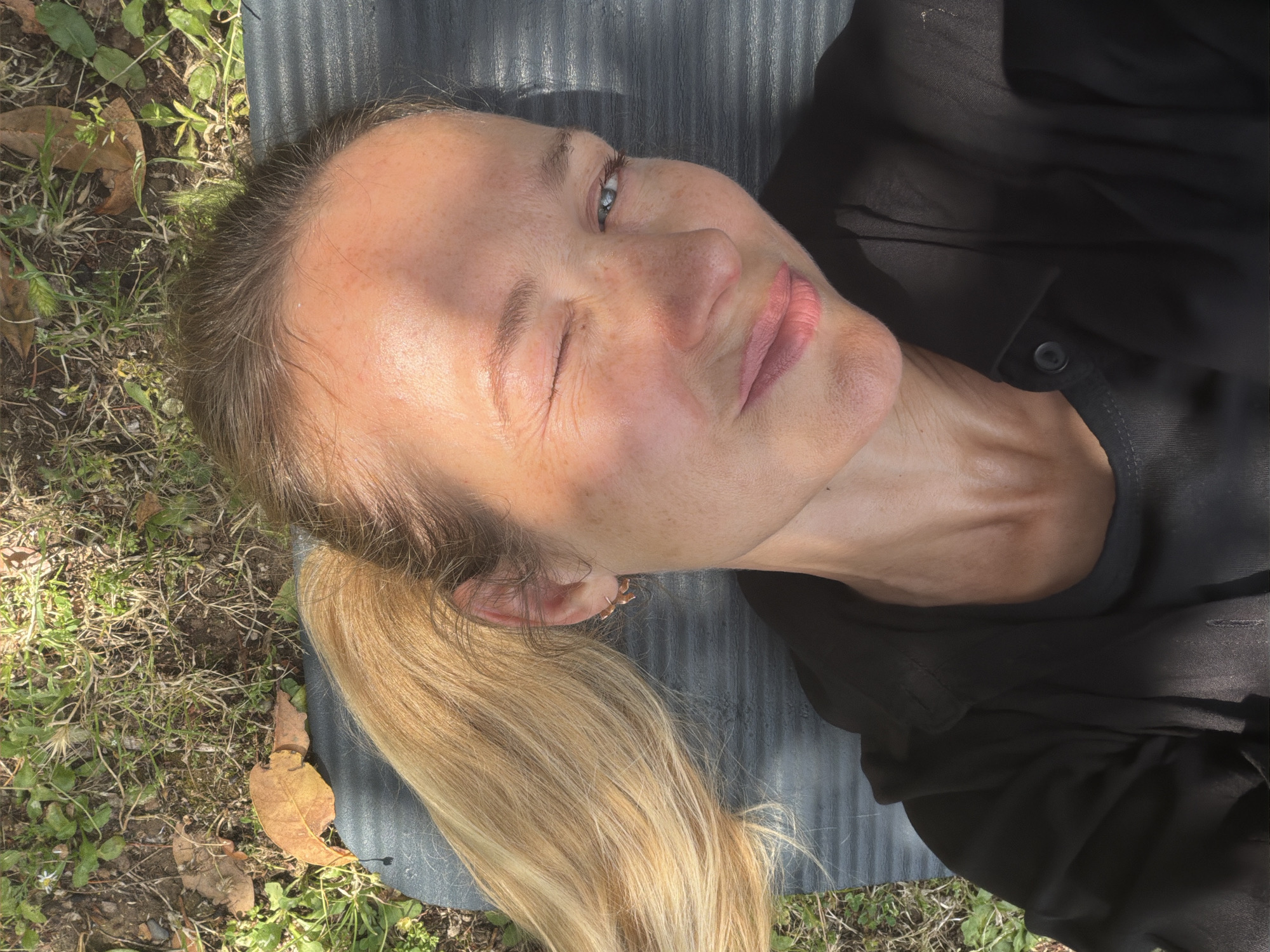 A blonde woman lies on her back on a dark yoga mat in a grassy, outdoor area, possibly a park or yard. She is wearing a black top and has her hair in a ponytail to her left. The woman is looking up at the camera and winking, with a slight smile on her face. Sunlight casts shadows across her face and shoulders. The background consists of green grass, dirt, and fallen leaves, suggesting an autumn or dry season. There are no visible buildings or urban structures to indicate a specific location within Budva, Montenegro.