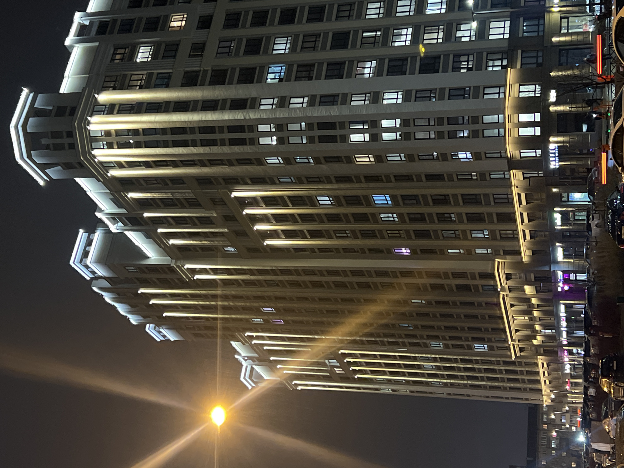 The image depicts a tall, multi-story building at night. The building is illuminated with horizontal bands of light. Windows punctuate the façade in a regular grid pattern. A street lamp shines in the lower left corner of the image. Cars are parked along the right side of the building. The location is Yekaterinburg, Russia.