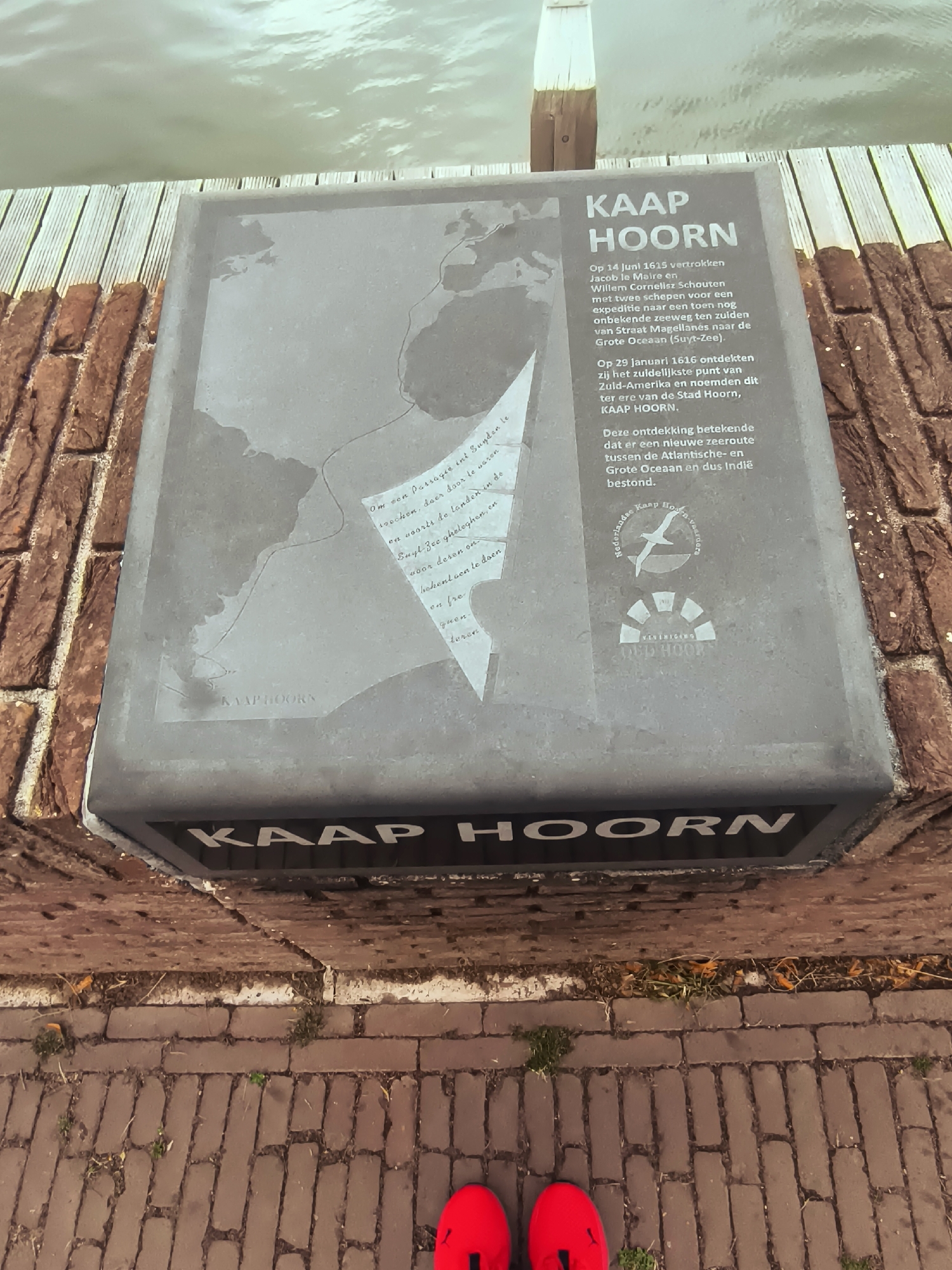 The image shows a plaque dedicated to "KAAP HOORN" (Cape Horn) located on a brick wall in the city of Hoorn, Netherlands. The plaque is mounted on a stone base and features a map, text in Dutch, and a logo. The text details the voyage of Jacob le Maire and Willem Cornelis Schouten in 1615, who discovered the southernmost point of South America and named it "Stad Hoorn, KAAP HOORN." It also mentions that this discovery led to a new sea route between the Atlantic and Indian Oceans. The environment appears to be outdoors, with a body of water and wooden decking visible in the background, suggesting a waterfront location. The foreground shows a brick walkway, and a pair of red shoes is visible at the bottom of the frame.