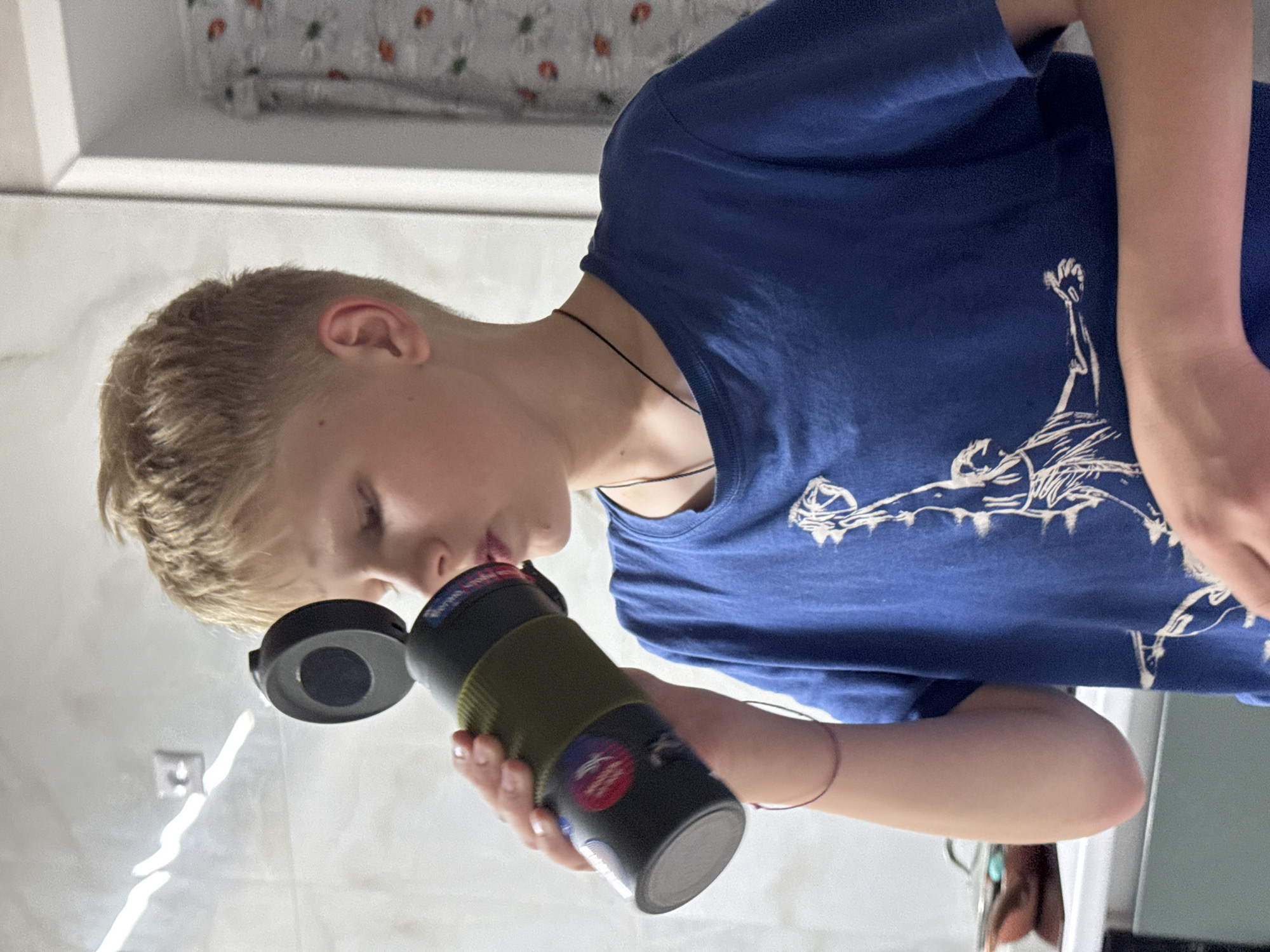 A fair-skinned adolescent is drinking from a black and green water bottle with stickers. He is wearing a blue t-shirt with a white design. The bottle cap is open and lying to the left. He appears to be in a bathroom with white tiled walls. The location is Ферма, Russia.