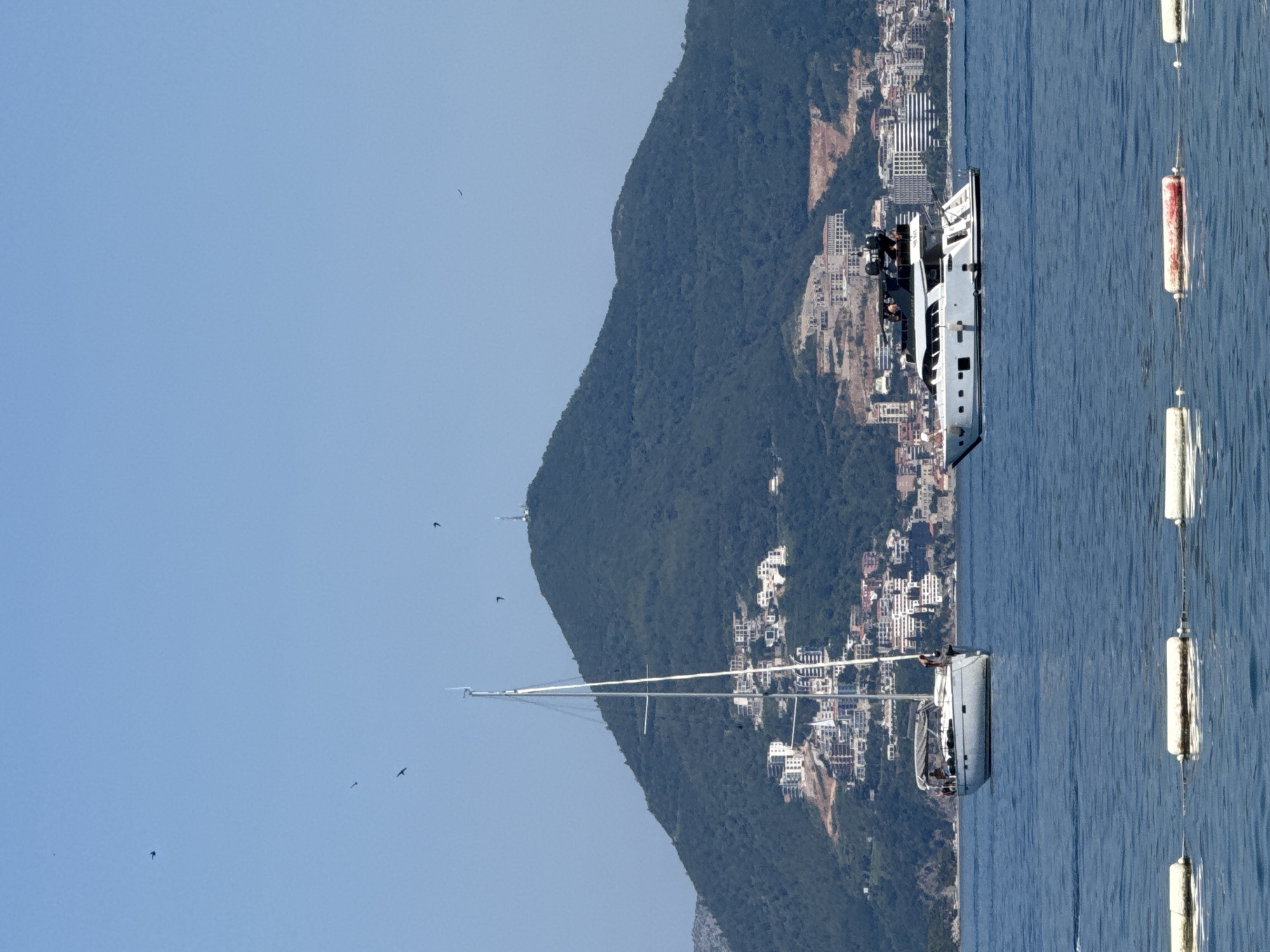 The image depicts a sunny day scene at sea, likely in or near Budva, Montenegro, with a large, verdant mountain dominating the background. A town, characterized by white buildings, is built on the lower slopes of the mountain. Two boats are visible in the foreground on the blue water: a white sailboat with its mast and rigging prominently displayed, and a sleek white motor yacht with black accents and several people visible on deck. In the far distance, other buildings and structures can be seen along the coastline. Several buoys, some white and one red and white, float on the water in the immediate foreground, marking a boundary or area. The sky is clear and blue, with a few dark specks that appear to be birds in flight.