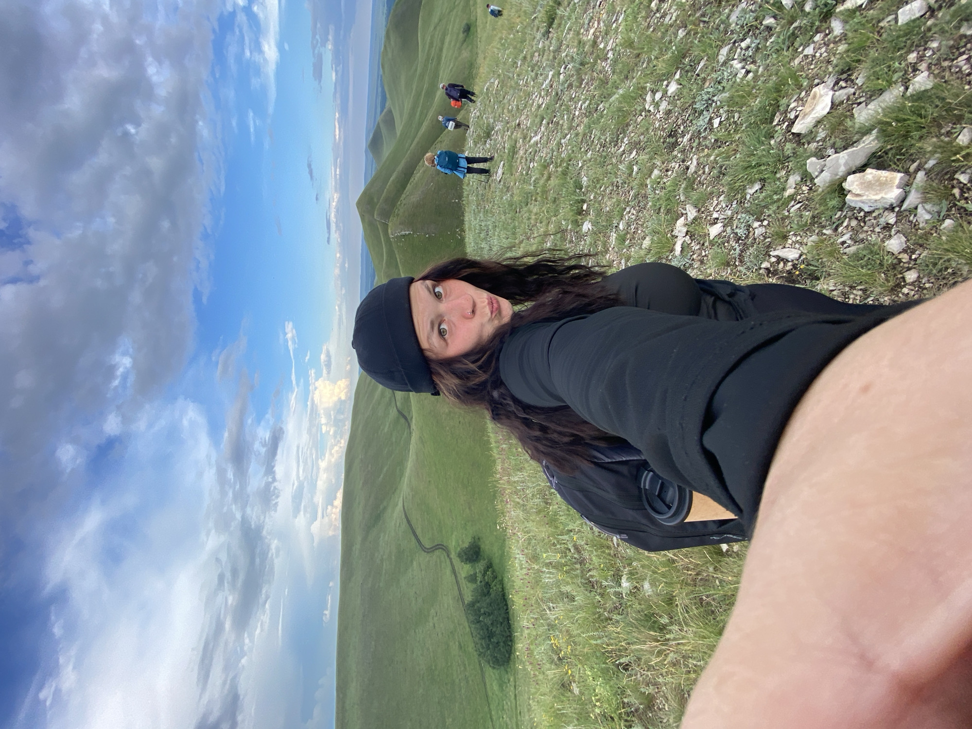 This is a portrait-oriented, full-frame photograph taken from the perspective of a person with long, dark, wavy hair and a black beanie, looking directly at the camera. They are wearing a black top and have a backpack on. Their facial expression is playful, with puckered lips and widened eyes.
In the background, there are several other people hiking on a grassy, hilly landscape under a partly cloudy sky. The terrain is characterized by rolling green hills with a winding path visible in the distance, and scattered rocks and vegetation in the foreground. The lighting suggests it is daytime, likely late afternoon or early evening, given the soft light and the dynamic cloud formations. The location is identified as Verkhneozernoe, Russia.
The other hikers appear to be dressed for outdoor activity, with some wearing backpacks and jackets. One person in the middleground is wearing a blue jacket and backpack, another in a purple jacket, and further up the hill, two more figures are visible. The overall scene depicts a group of people engaged in hiking or trekking in a scenic, mountainous or hilly environment. No text is visible in the image.