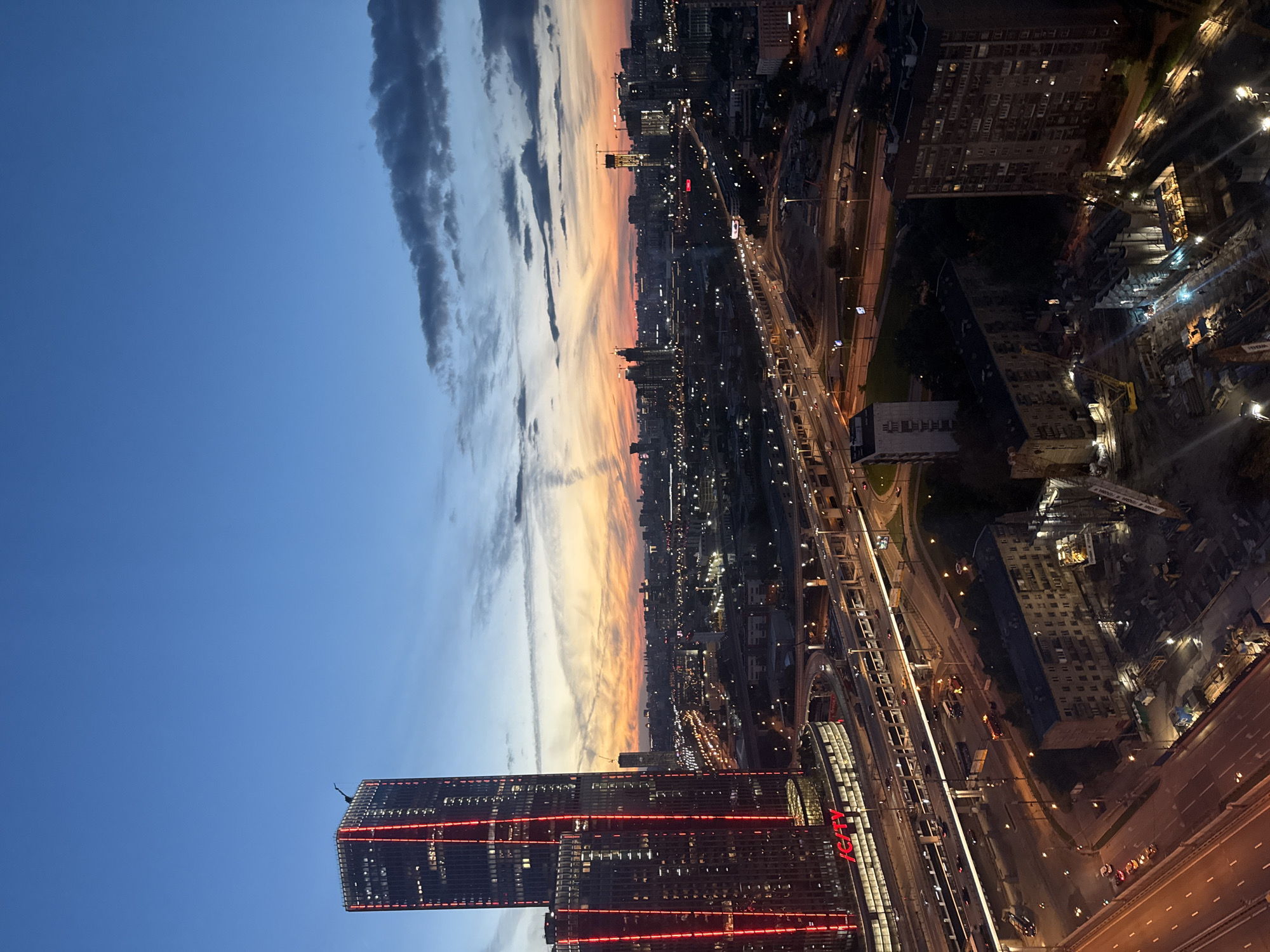 An aerial shot captures a cityscape at dusk, with a clear blue sky transitioning into an orange and pink sunset horizon. A prominent skyscraper with red vertical lighting dominates the left side of the frame, casting a glow on adjacent buildings. A complex network of highways and roads is visible below, illuminated by streetlights and car headlights. In the distance, a sprawling urban landscape stretches out, dotted with more buildings and construction cranes. The overall scene depicts a modern city undergoing development, with both established structures and ongoing projects visible.