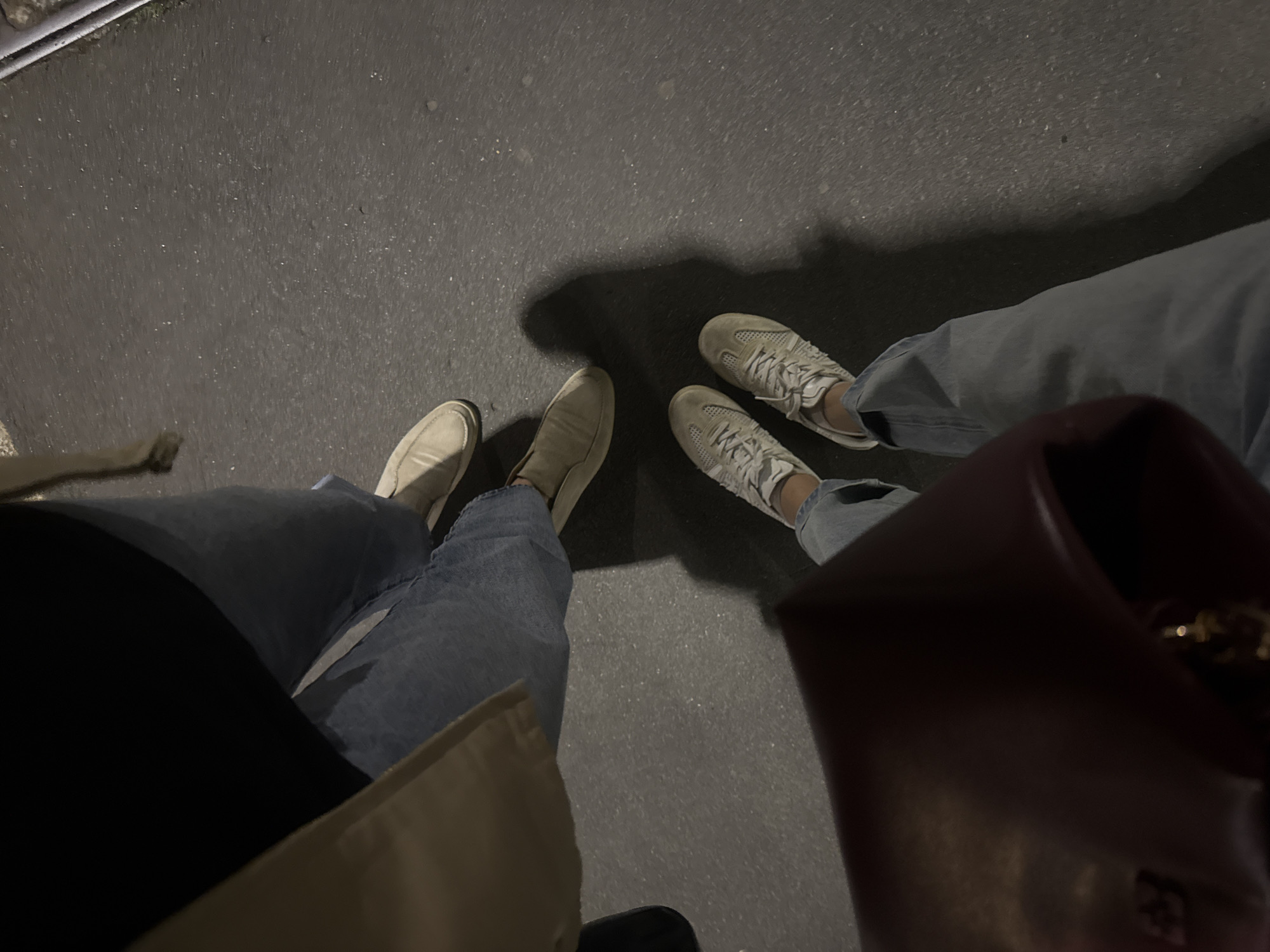 The image depicts a street scene at night, likely in an urban area. Two pairs of feet are visible, belonging to two individuals standing on a paved surface, possibly a sidewalk or pedestrian area. The person on the left is wearing light-colored loafers, and blue jeans, with parts of a black jacket and a beige coat visible. The person on the right is wearing light-colored sneakers and blue jeans. A dark-colored bag is visible on the right side of the image, partially obscuring the view. The pavement is dark gray. The location is unknown, so I cannot confirm it is in Milan.
