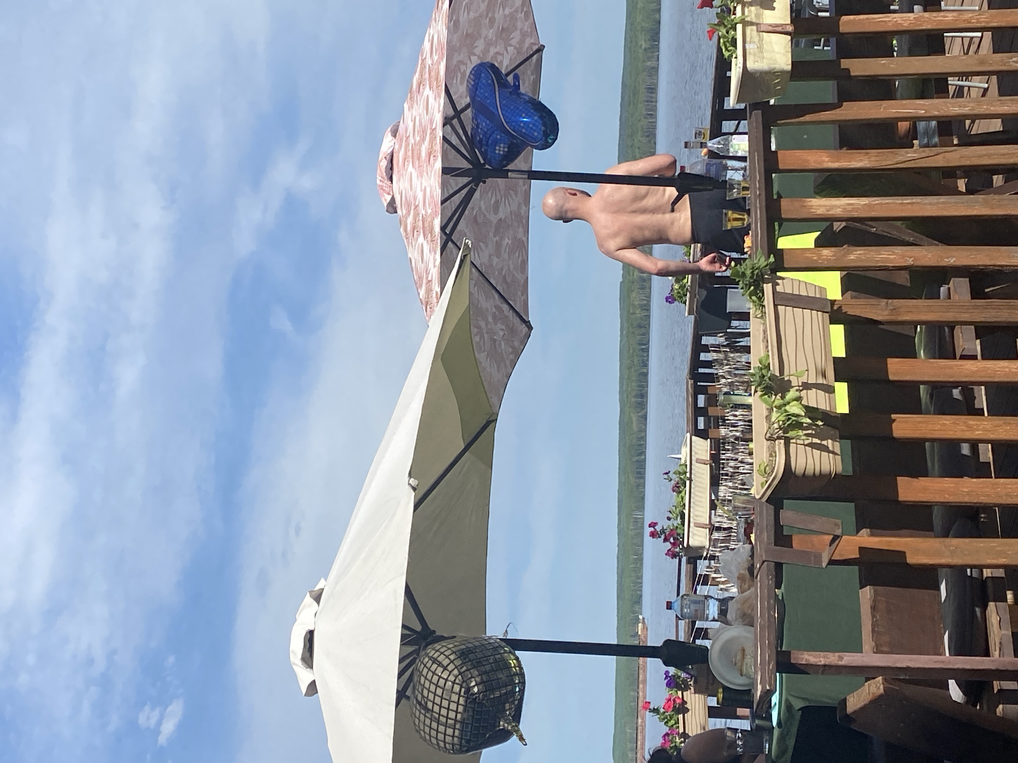 Scene: An outdoor terrace overlooking a body of water, possibly the Kama River in Chaikovsky, Russia.
Objects:
- Two large umbrellas: one primarily white with a disco ball hanging from it, and another with a floral pattern and a blue balloon attached.
- A wooden railing surrounds the terrace.
- Planters with flowers are situated on the railing.
- Bar area with bottles, glasses, and other bar accessories.
People:
- A bald man is standing near the bar, shirtless and wearing dark shorts.
Environment:
- The sky is blue with scattered white clouds.
- A body of water (possibly the Kama River) is visible in the background.
- Green trees line the opposite bank of the water.
Actions/Interactions: The man is standing near the bar, possibly preparing a drink. The scene suggests a relaxed, outdoor setting.