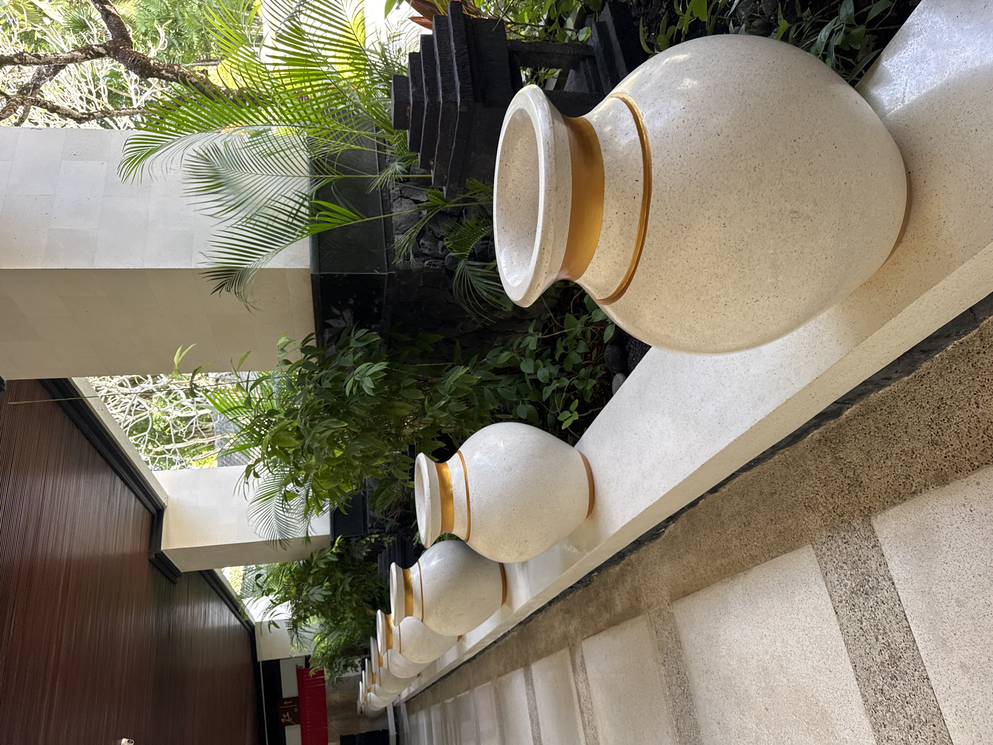 The image depicts a row of decorative, round, cream-colored urns with gold-colored bands around their necks, resting on a cream-colored wall. The urns decrease in size as they recede into the distance. The background includes green foliage, palm fronds, and architectural elements like a tiered water feature constructed of dark stone. To the left, a dark brown wooden wall is visible. The scene is set in Kuta Selatan, Indonesia. The image is taken at a low angle looking up, and the overall scene conveys a sense of tranquility and outdoor decor.
