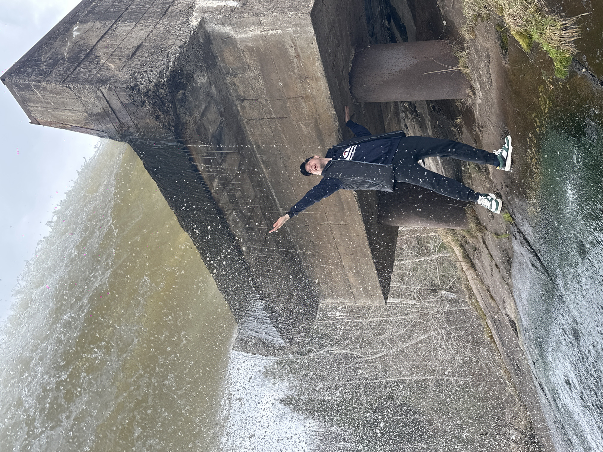 A rotated medium shot depicts a man standing under a concrete structure near a waterfall. The man, wearing a black cap, black jacket, and patterned pants, is positioned with his arms outstretched. His feet are clad in white and green sneakers. The concrete structure looms above him, with water cascading down one side. Vegetation grows along the edge of the structure. The water is murky. The scene is captured during daylight hours with overcast lighting. This image was taken near the Votkinsk Hydroelectric Station in the city of Chaikovsky, Russia.