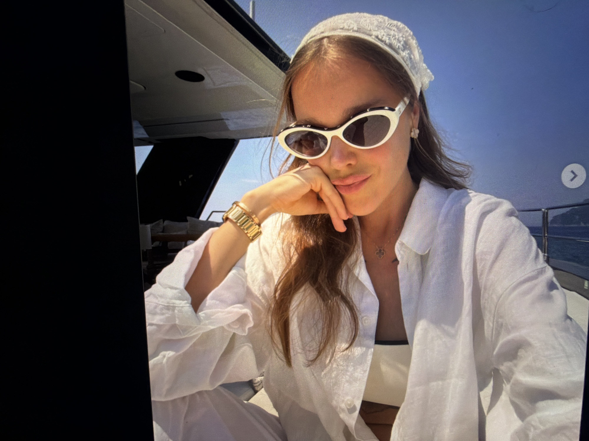 A woman with brown hair is wearing a white bandana, white cat-eye sunglasses, and a white linen shirt. She has a gold bracelet on her left wrist and a necklace with a small clover pendant. She is resting her chin on her hand and looking at the camera. The background is a bright blue sky with a hint of the ocean and a boat. The image appears to be a selfie taken on a boat. The location in the city of Falenty Nowe, Poland is not identifiable from the image.
