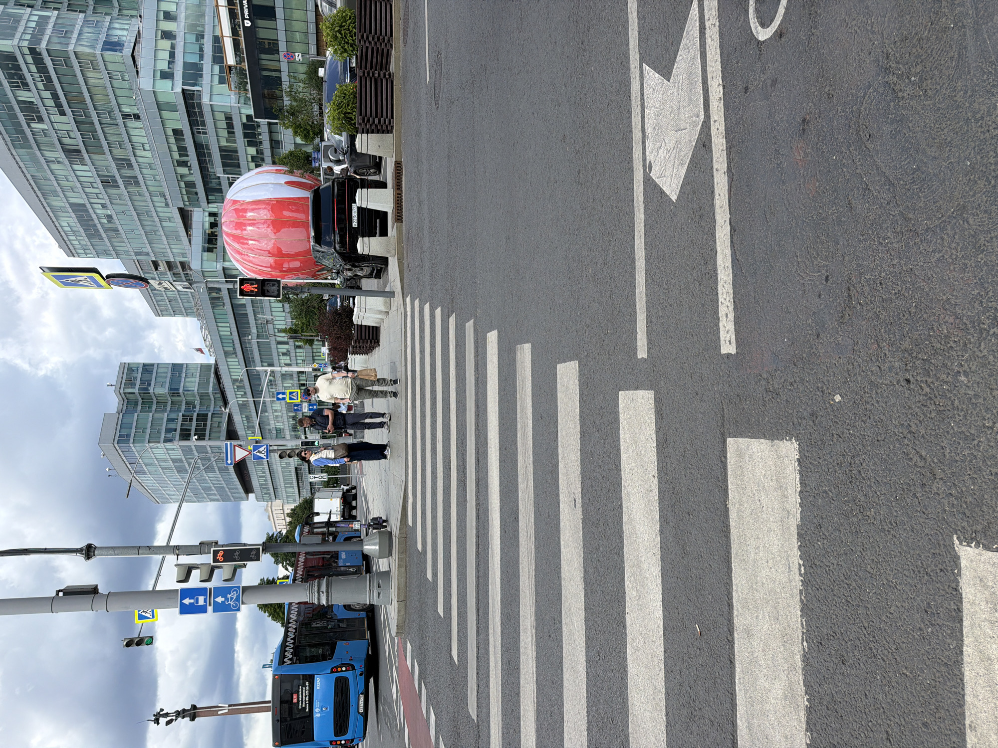 The image captures a street scene in Moscow, Russia. In the foreground, a pedestrian crossing with prominent white stripes stretches across the asphalt. To the right of the crossing, a large white arrow is painted on the road, indicating a direction of travel. Behind the pedestrian crossing, several people are visible, appearing to be walking or waiting. A blue bus is parked on the left side of the street, with some text visible on its rear.
In the middle ground, traffic lights and street signs are mounted on poles. One traffic light shows a red signal for vehicles, while another appears to be displaying a green light for pedestrians or cyclists. The background is dominated by modern, multi-story buildings with glass facades, suggesting an urban environment. A large, red and white balloon-like object is prominently placed near the buildings. The sky is overcast, indicating a cloudy day. The overall scene depicts a typical urban street with pedestrian and vehicular traffic, set against a backdrop of contemporary architecture.