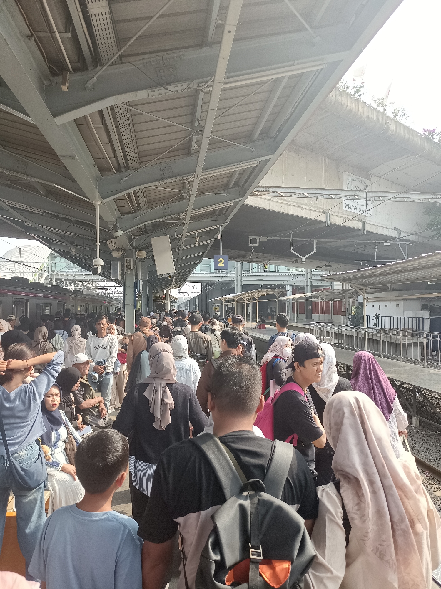 The image shows a crowded train platform at Wijaya Karya station in Central Jakarta, Indonesia. Many people are standing, waiting, and some are walking. The platform is covered by a large, grey metal roof supported by pillars. Trains are visible on the tracks to the left of the image. The atmosphere appears to be busy, with a large number of commuters. The lighting suggests it is daytime, with the sun partially obscured by the roof structure, creating bright highlights. There are signs and infrastructure typical of a train station, including signage with the station name and route numbers.