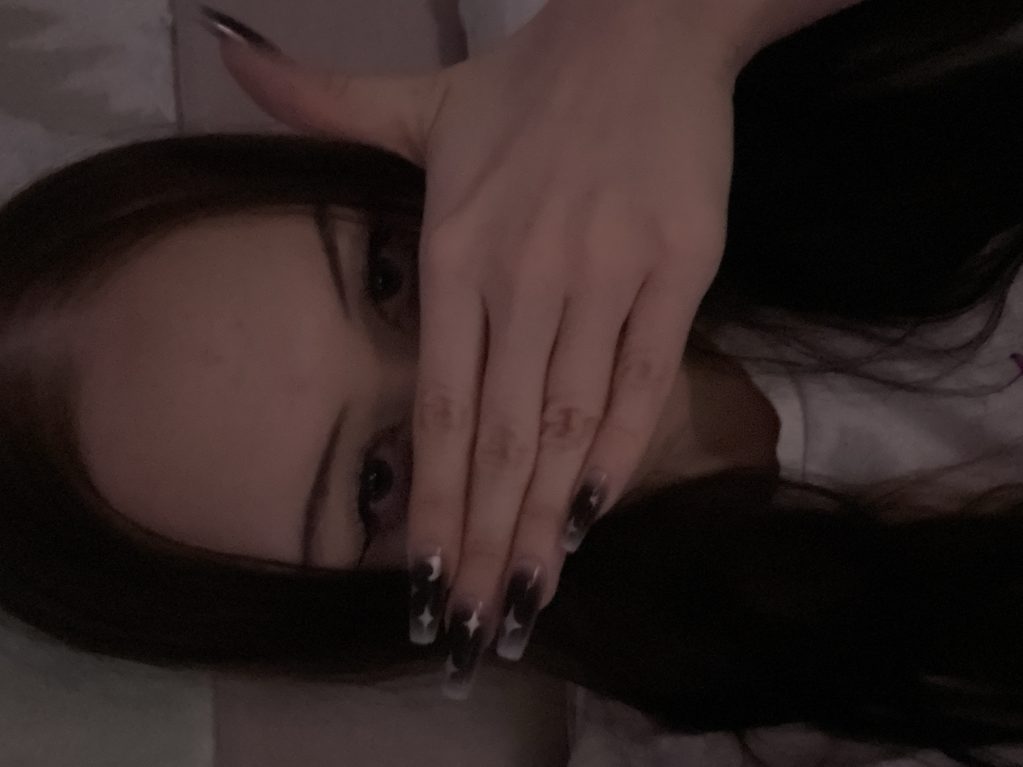 A young woman with long, dark hair and dark eyeliner is covering the lower half of her face with her right hand. Her nails are long and appear to be decorated with dark tips and star-shaped designs. She is wearing a white t-shirt. The background is indistinct and appears to be a dimly lit room with a light-colored surface behind her. There is no visible indication of the city of Votkinsk, Russia.