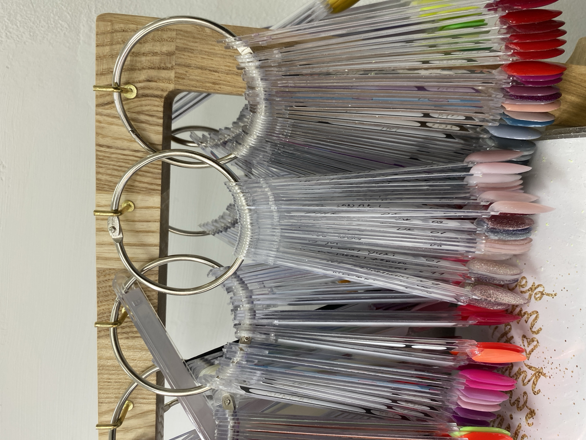 The image displays a comprehensive collection of nail polish color swatches, likely intended for client selection in a beauty or nail salon setting in Chaikovsky, Russia. No people are visible.
The primary subjects are numerous clear plastic nail tips, each painted with a distinct nail polish color or finish. These tips are organized into several bundles, fanning out from large silver metal rings. Each ring holds a substantial quantity of swatches. These rings are, in turn, attached to a light-colored wooden board or panel, which exhibits a visible wood grain. Small, brass-colored fixtures secure the rings to the wooden board.
The swatches themselves showcase a wide spectrum of colors, including vibrant reds, various shades of pink and nude, cool blues and greys, rich purples, and bright oranges and greens. Many swatches feature a glossy finish, while others exhibit glitter, shimmer, or matte effects. The clear plastic sticks extend beyond the painted tips, often bearing handwritten labels.
The implied activity is the selection of a nail polish color by a customer or nail technician. The methodical arrangement of the swatches suggests a well-organized system for presentation and choice. The overall lighting appears bright, possibly indicating an indoor, well-lit environment during the daytime.
Visible text on the clear plastic sticks includes various alphanumeric codes and names, such as "09", "01", "Yuki", "Lall", "DR", "02", "Dior", and "GLAM", which are likely color codes, brand names, or specific shade identifiers. On a white surface to the right, a gold glittery script is partially visible, appearing decorative.