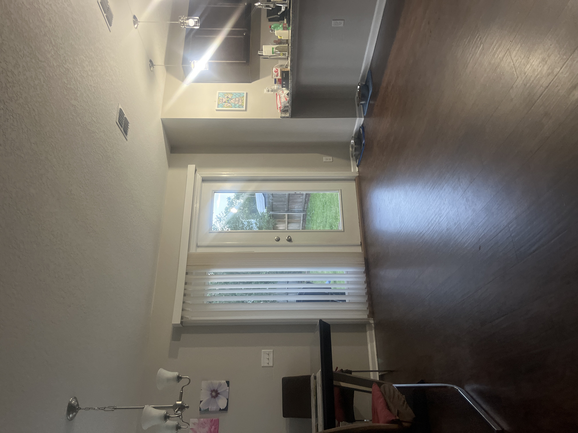 This is a full-shot, eye-level, indoor photograph of a dining and kitchen area in Jacksonville, United States of America. The room has dark wood flooring and light grey walls. In the foreground, a dark dining table with chrome legs and a dark chair with a red cushion are visible on the left. Two floral art pieces hang on the wall behind the table. A chandelier with three lights hangs from the ceiling above the dining area.
A sliding glass door with vertical blinds leads to a backyard with a green lawn and a wooden fence. Two ceiling vents are visible in the ceiling. In the background, a kitchen counter with dark cabinets above and to the right is present. Several items are on the counter, including bottles, containers, and a coffee maker. Two dog bowls on blue mats are placed on the floor near the kitchen counter. The lighting appears to be artificial, with overhead lights and light coming from the backyard.
