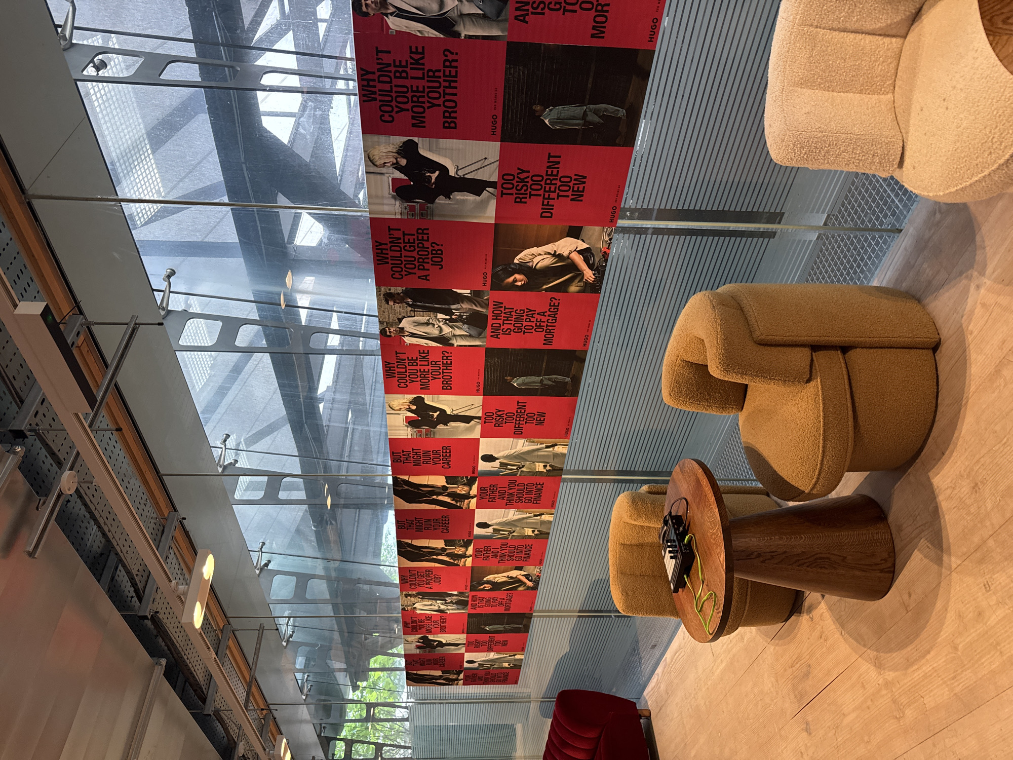 The image shows an interior space with a partial view of a glazed roof structure and several pieces of furniture. A series of red promotional posters are suspended in front of the glazing, repeating slogans and images associated with a clothing brand, HUGO. The text on the posters includes phrases like "WHY COULDN'T YOU BE MORE LIKE YOUR BROTHER?" and "TOO RISKY TOO DIFFERENT TOO NEW".
The furniture consists of plush, rounded armchairs in various shades of tan and cream, arranged to create small seating areas. A round, dark wood side table with a unique trumpet-shaped base sits between two armchairs, holding a small black electronic device with a green cable plugged into it. A red velvet sofa is also partially visible in the lower portion of the image.
The location seems to be a modern, possibly temporary, installation or showroom, though it is difficult to determine the specific location in London, United Kingdom from this image. The overall impression is a contemporary, stylishly designed space with promotional branding and furniture.
