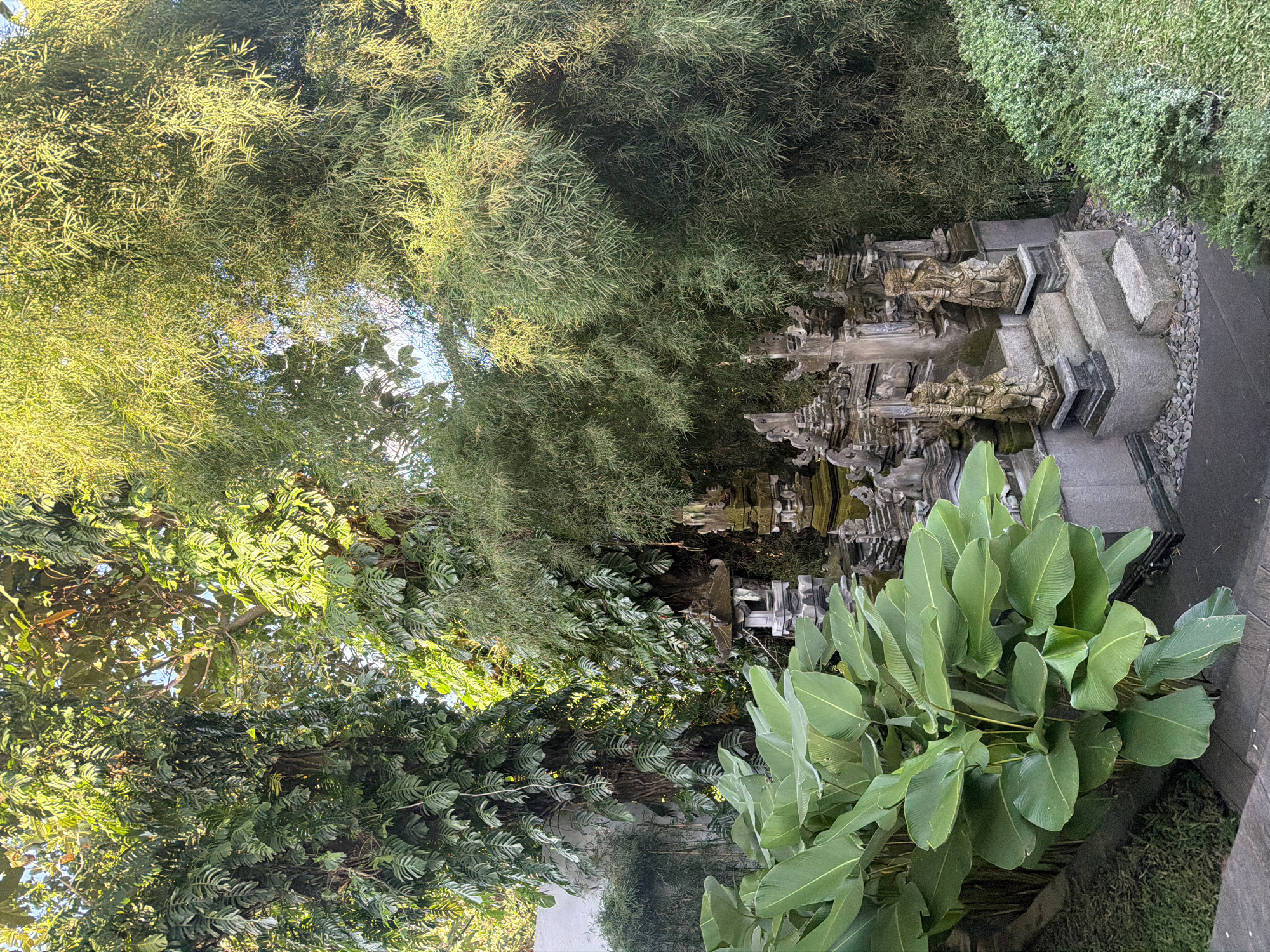 This image depicts a garden scene, likely in Mas, Indonesia, known for its wood carving artistry. The scene features lush vegetation, including bamboo, large leaf plants, and other tropical foliage. A series of tiered stone structures with sculpted figures are visible amongst the plants, suggesting a water feature or shrine within the garden. The lighting indicates daylight hours, with dappled sunlight filtering through the trees. The arrangement suggests a deliberate landscaping effort to create a tranquil and aesthetically pleasing environment. No people are visible.