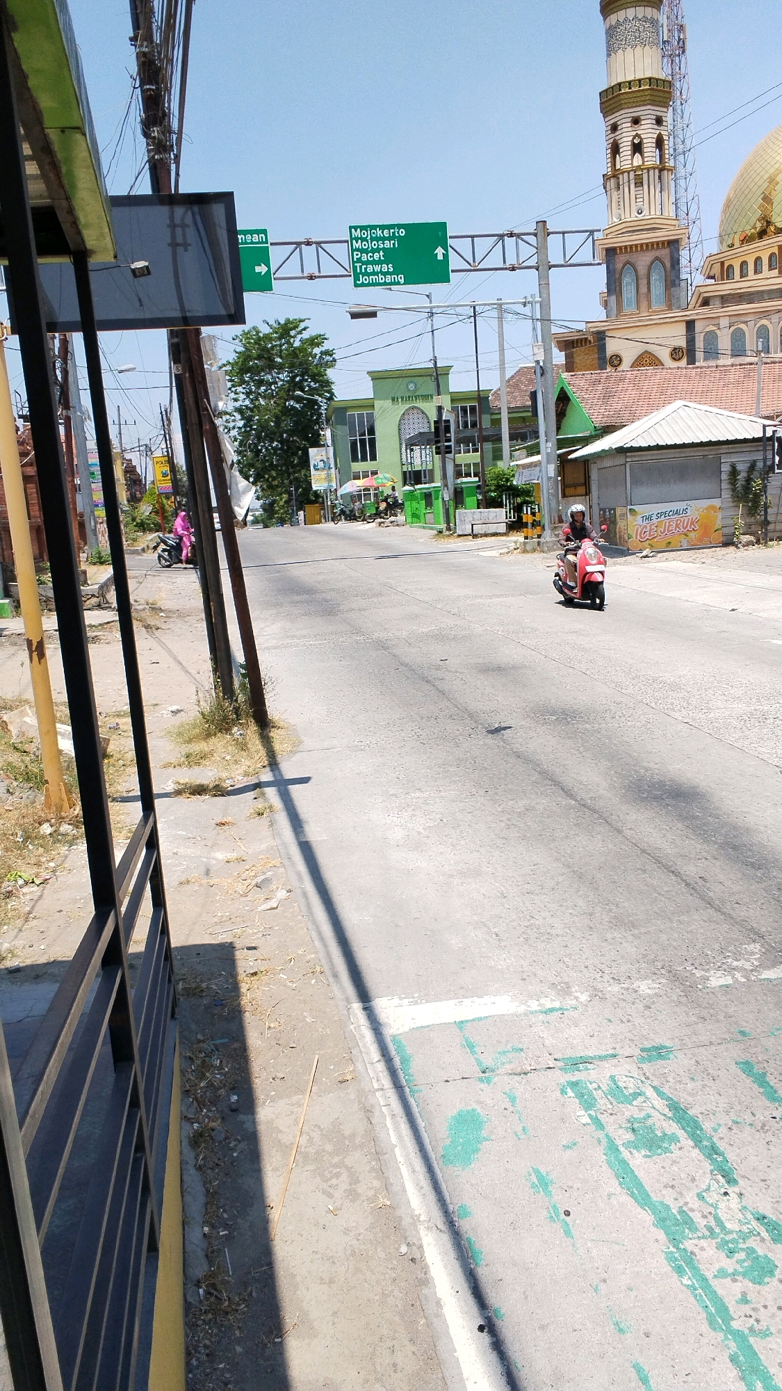 The image shows a street scene in Pulorejo, Indonesia, with a large mosque prominently visible on the right. A green directional signpost indicates "Mojokerto, Mojosari, Pacet, Trawas, Jombang" and points straight ahead. On the left, a building with a green facade and a sign that reads "MA NURULHUDA" is visible. A man is riding a red scooter down the street, wearing a helmet. Another person in a pink outfit is riding a pink scooter further down the road on the left. The sky is clear and blue, and the sun is shining. The street is paved, and there are power lines overhead. The area appears to be a commercial or residential district with several buildings and structures.
